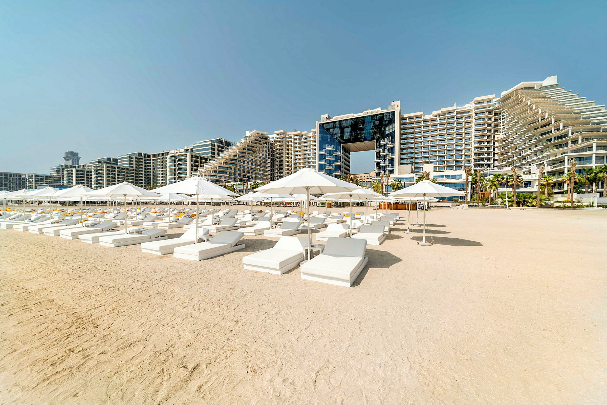 Solstolar och parasoller på en sandstrand med ett hotellkomplex i bakgrunden. (AI-genererad text)