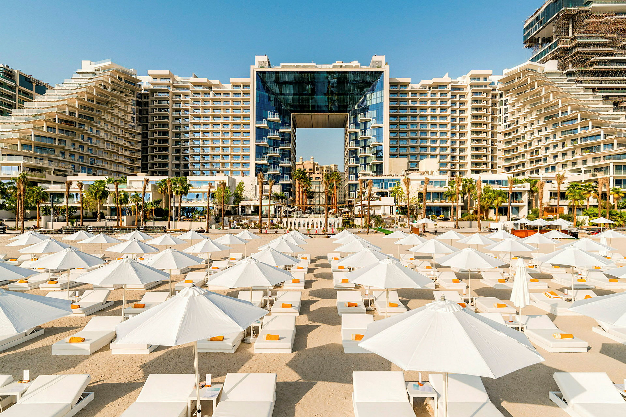 Hotellbyggnad med modern arkitektur, omgiven av palmer och framför en strand med vita parasoller och solstolar. (AI-genererad text)