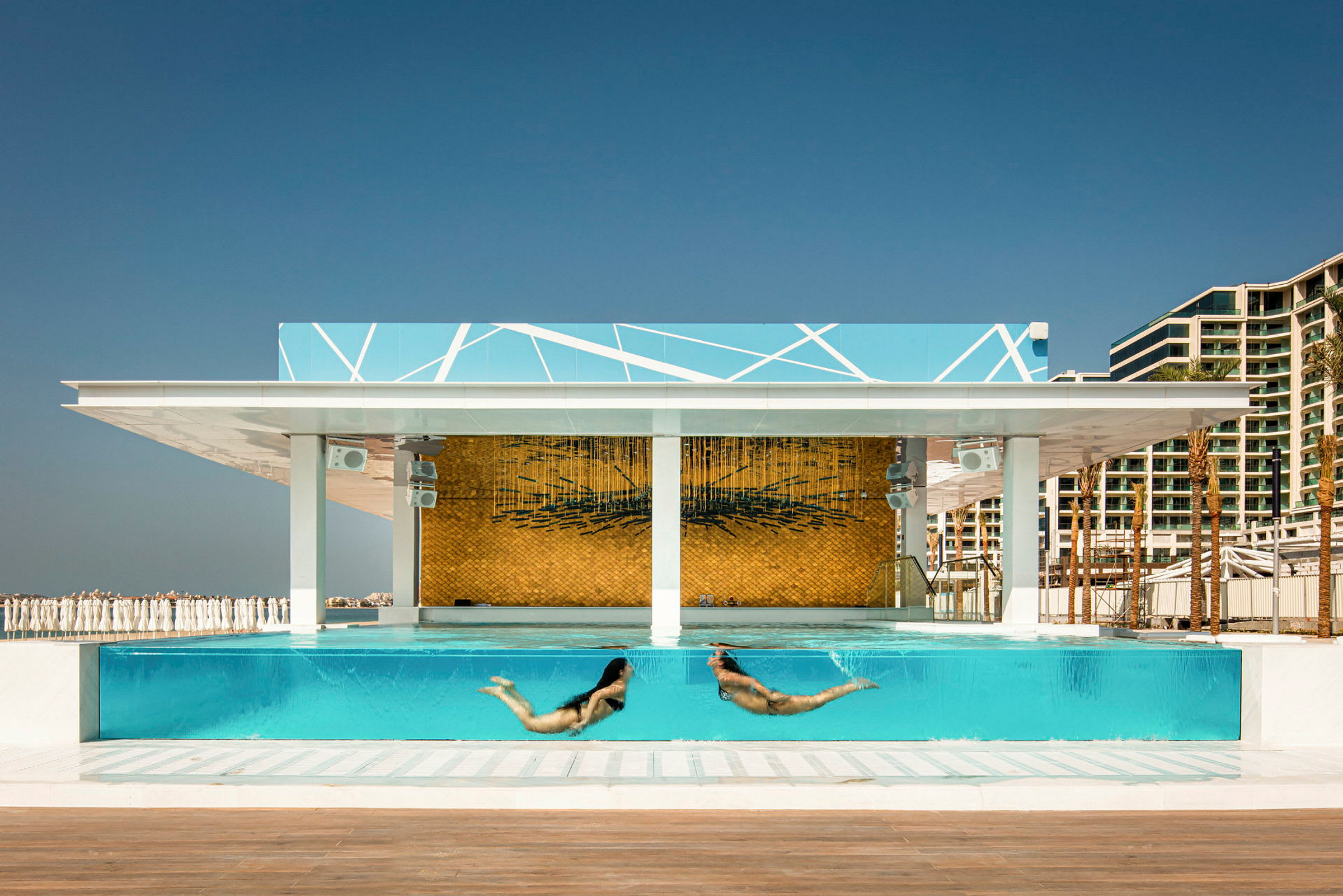 Två personer simmar i en pool med glasväggar. Bakgrunden visar modern arkitektur och en konstnärlig vägg. (AI-genererad text)