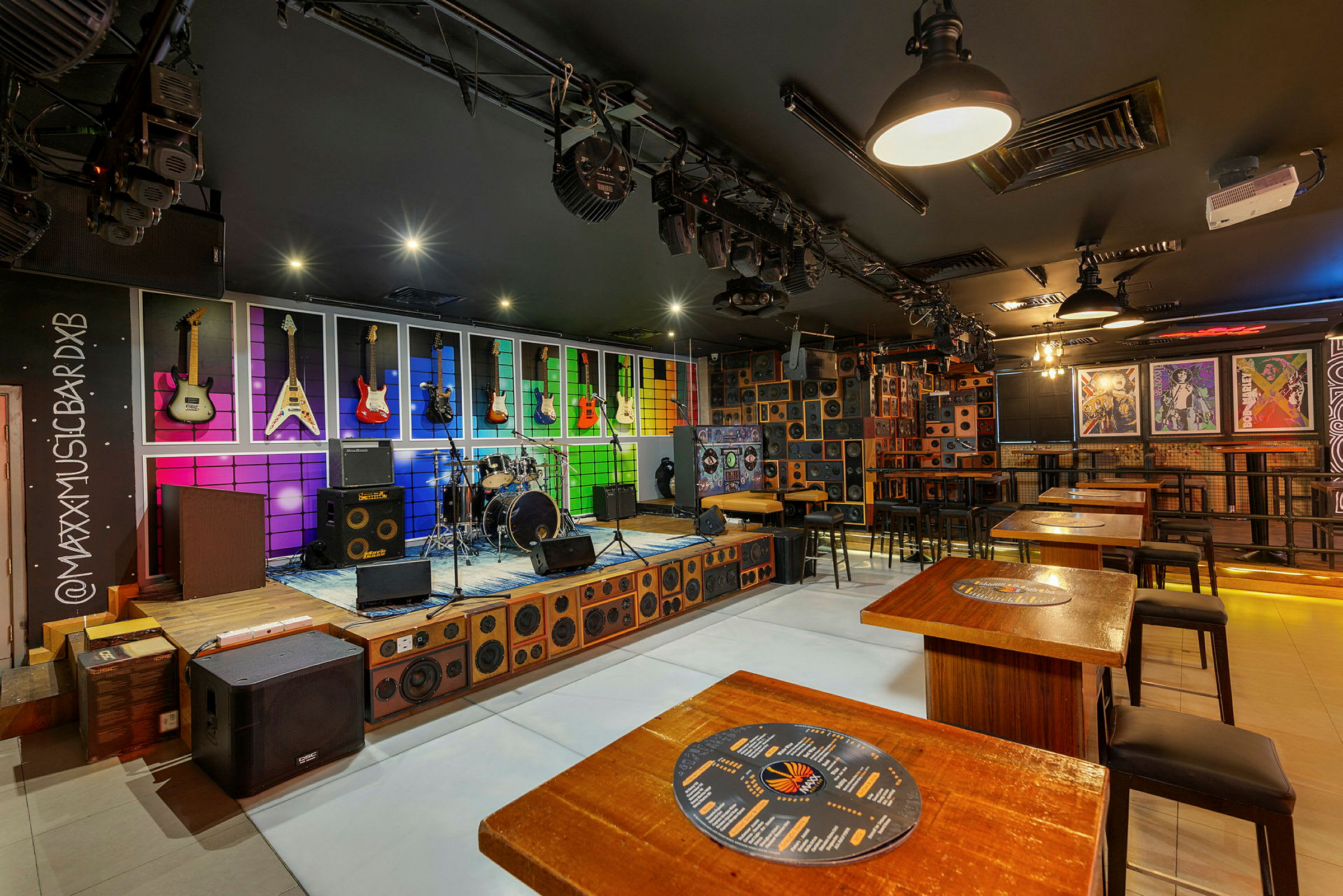 En interiör av en musikbar med färgglada gitarrer på väggen bakom trumset, högtalare och sittplatser. (AI-genererad text)