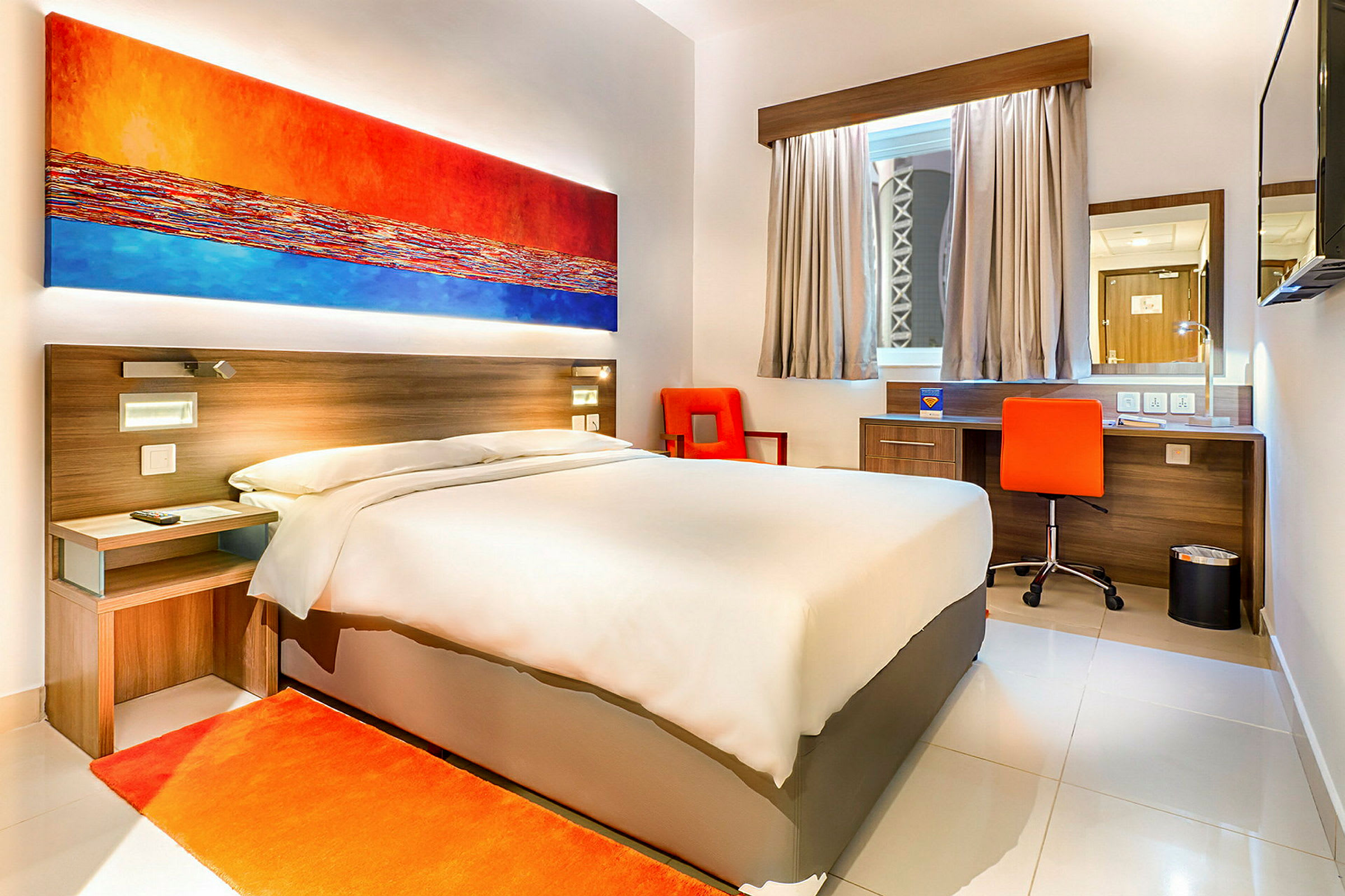 Ett modernt hotellrum med en dubbelsäng, färgstark väggkonst och ett skrivbord med en orange stol. (AI-genererad text)