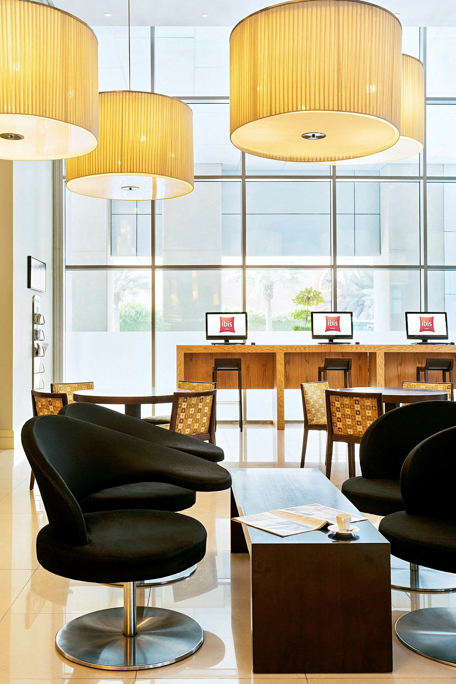 Ett hotellobbyområde med svarta fåtöljer, ett lågt bord och datorer på en barliknande disk framför stora fönster. (AI-genererad text)