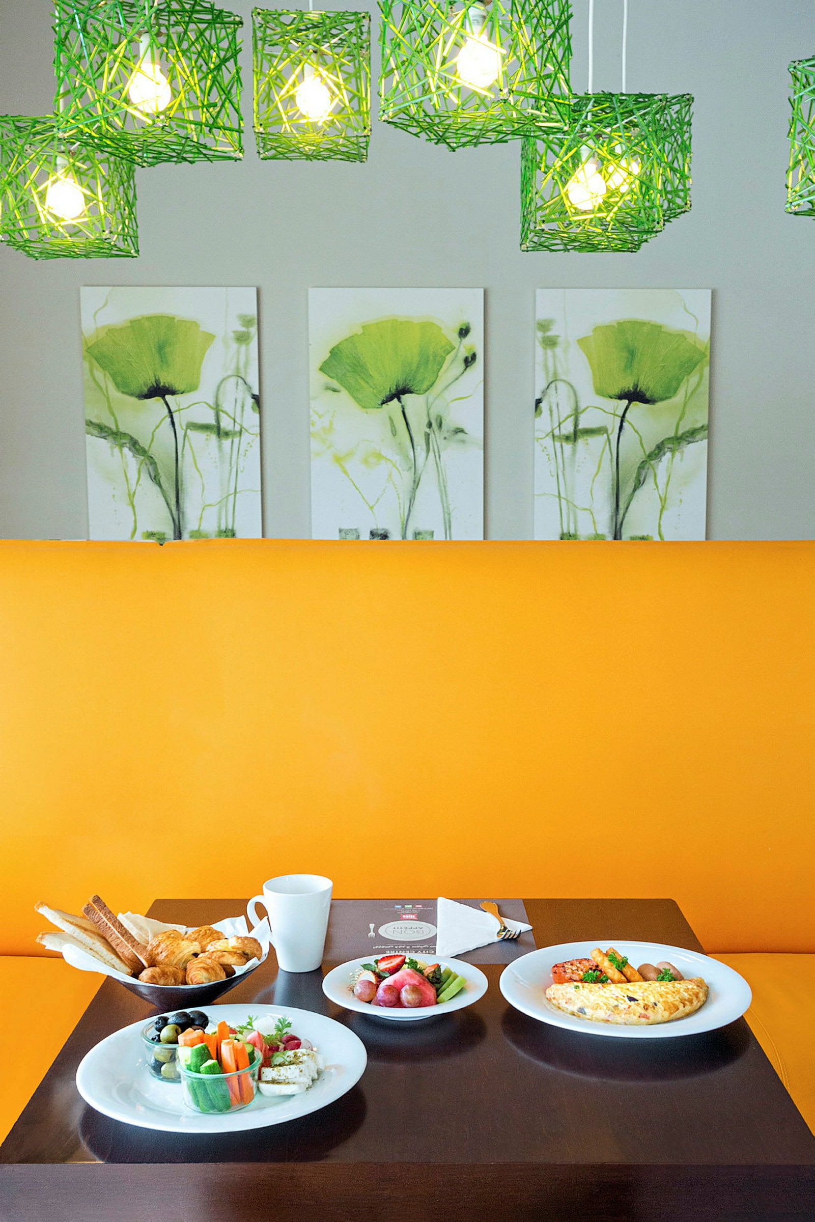 Ett bord fyllt med frukostmat, inklusive bröd, grönsaker och omelett, framför en gul sittplats med gröna lampor och blomkonst i bakgrunden. (AI-genererad text)