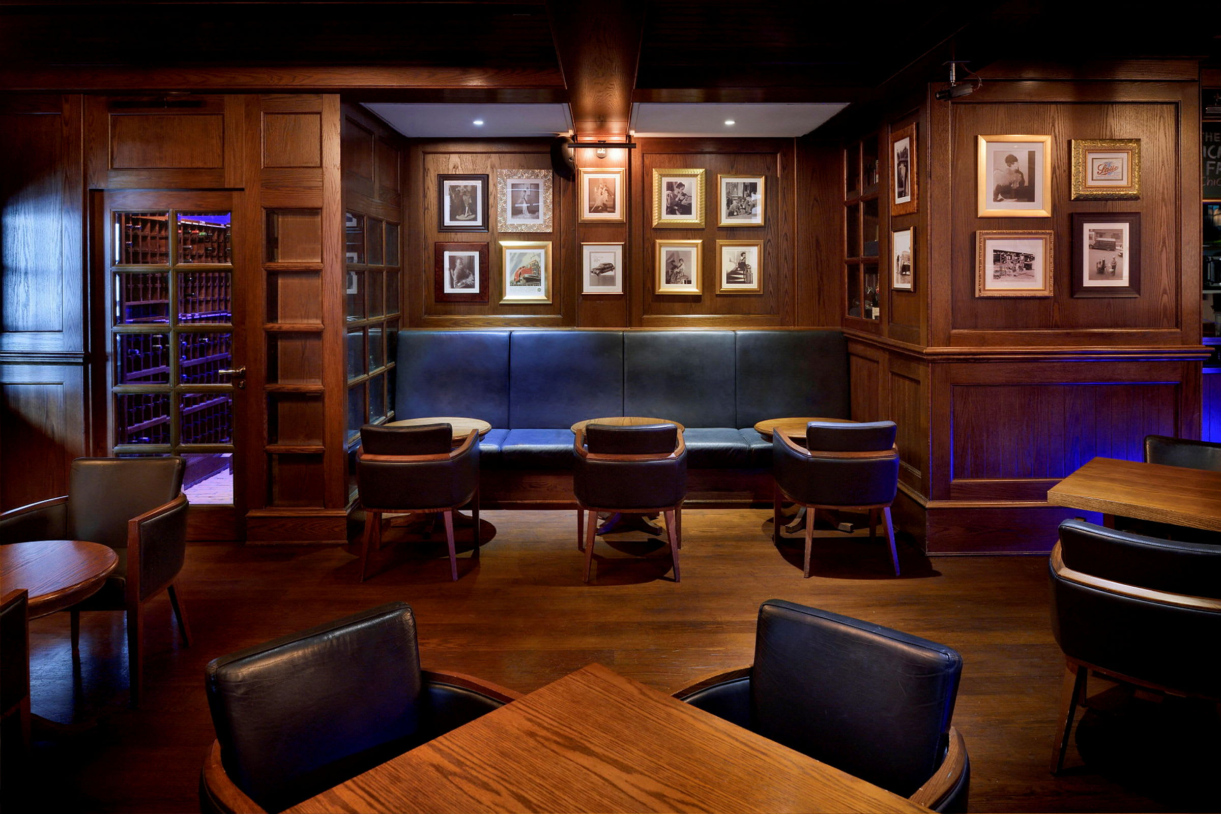 Ett rum inrett som en lounge eller bar med träpaneler, inramade bilder på väggen, stoppade bänkar och stolar samt små bord. (AI-genererad text)