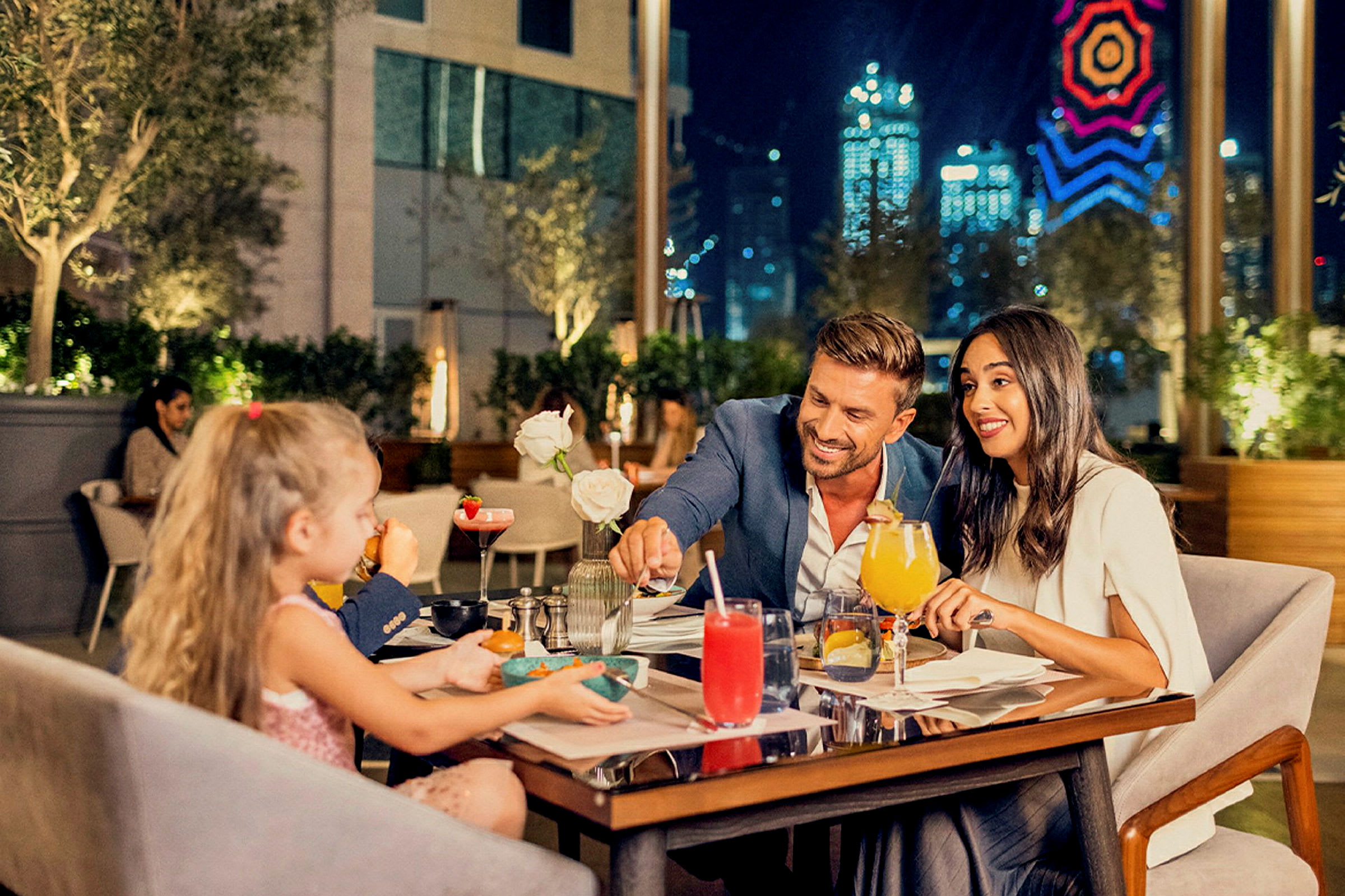 En middagsscen på en restaurang med uteservering. Bakgrunden visar modern arkitektur och skimrande stadsljus. (AI-genererad text)