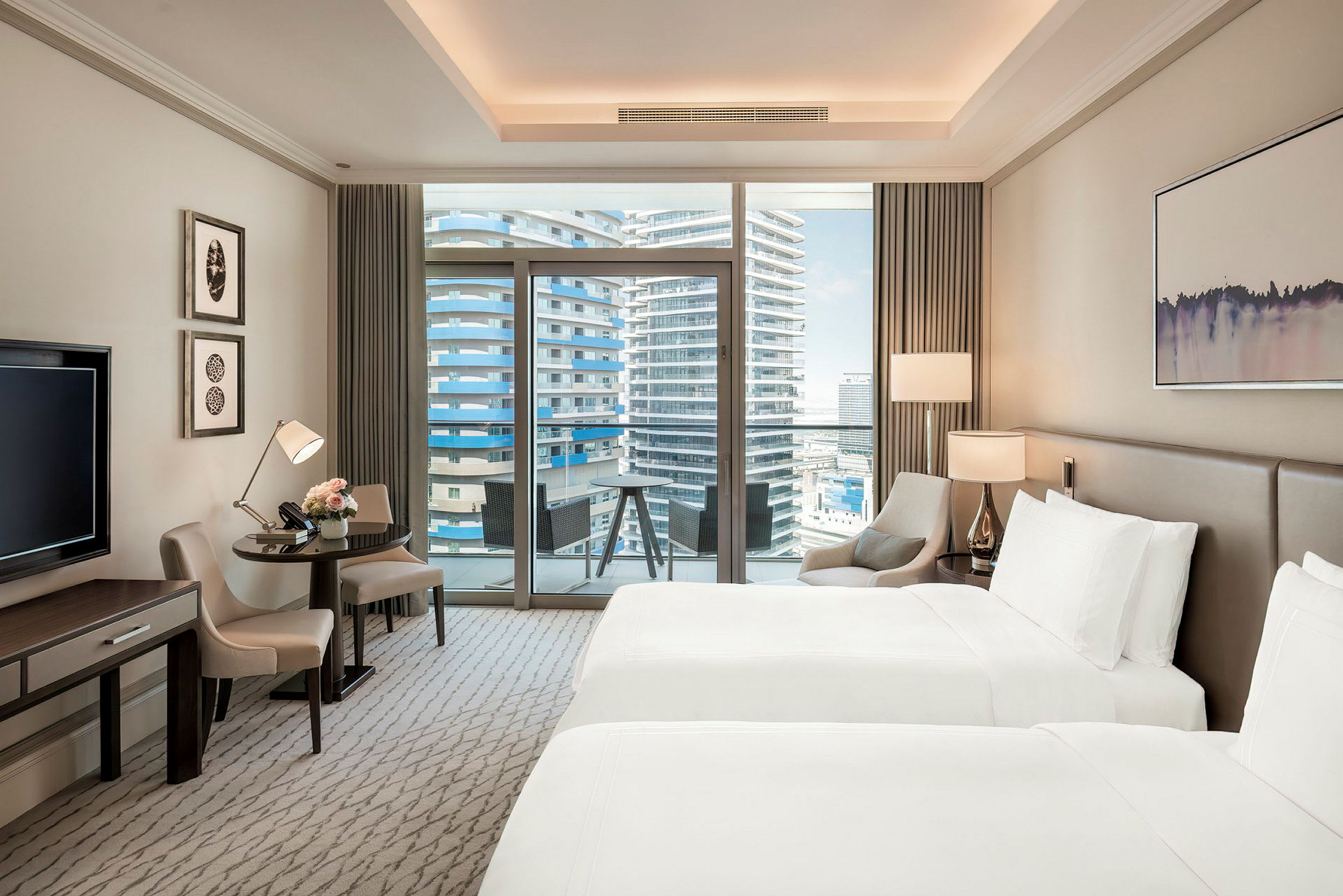 Ett modernt hotellrum med två sängar, ett skrivbord, ett litet bord och stolar, samt en balkong med utsikt över höga byggnader. (AI-genererad text)