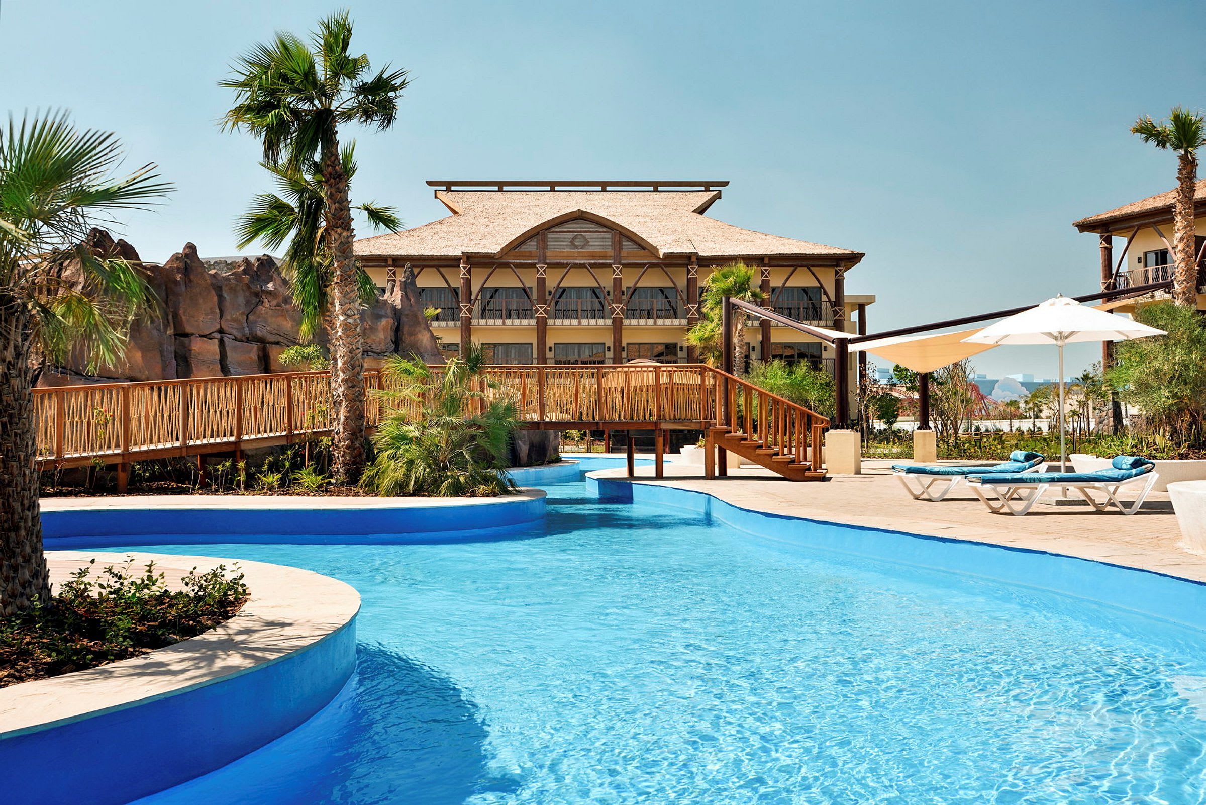 En pool med blått vatten och en träbro, omgiven av palmer och byggnader. (AI-genererad text)