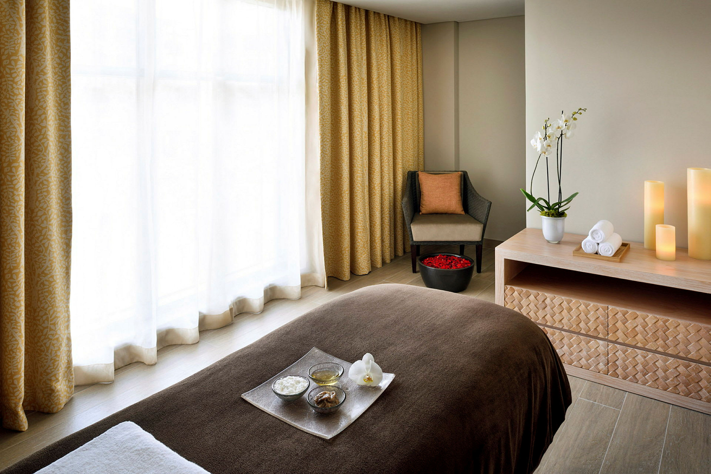 Ett spa-rum med massagebänk, guldfärgade gardiner, en sittplats med kudde, ett bord med handdukar och dekorativa ljus samt en skål med rosenblad. (AI-genererad text)