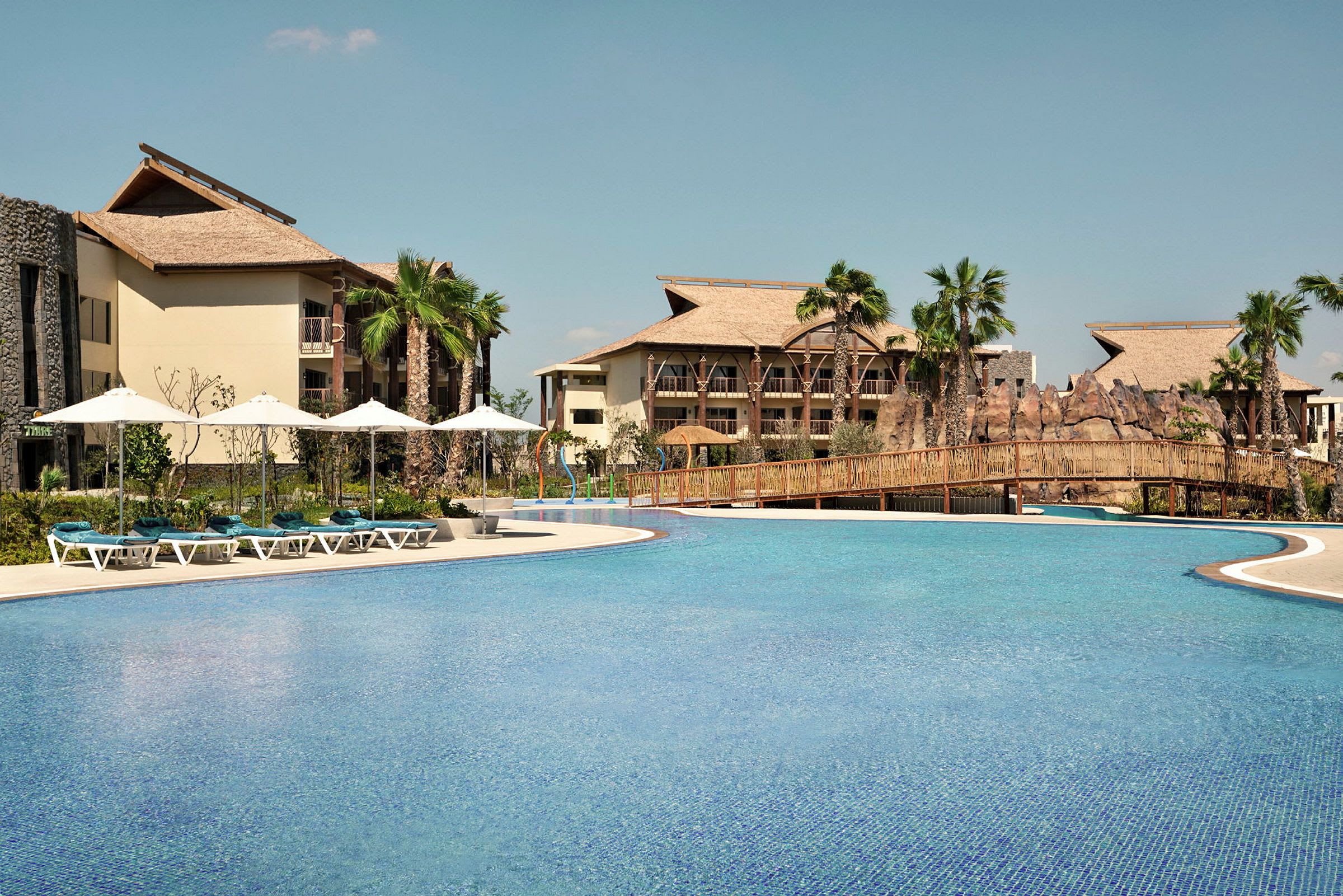 Stor pool med solstolar och parasoller i förgrunden. Hotellbyggnader med palmträd och en träbro över vattnet syns i bakgrunden. (AI-genererad text)