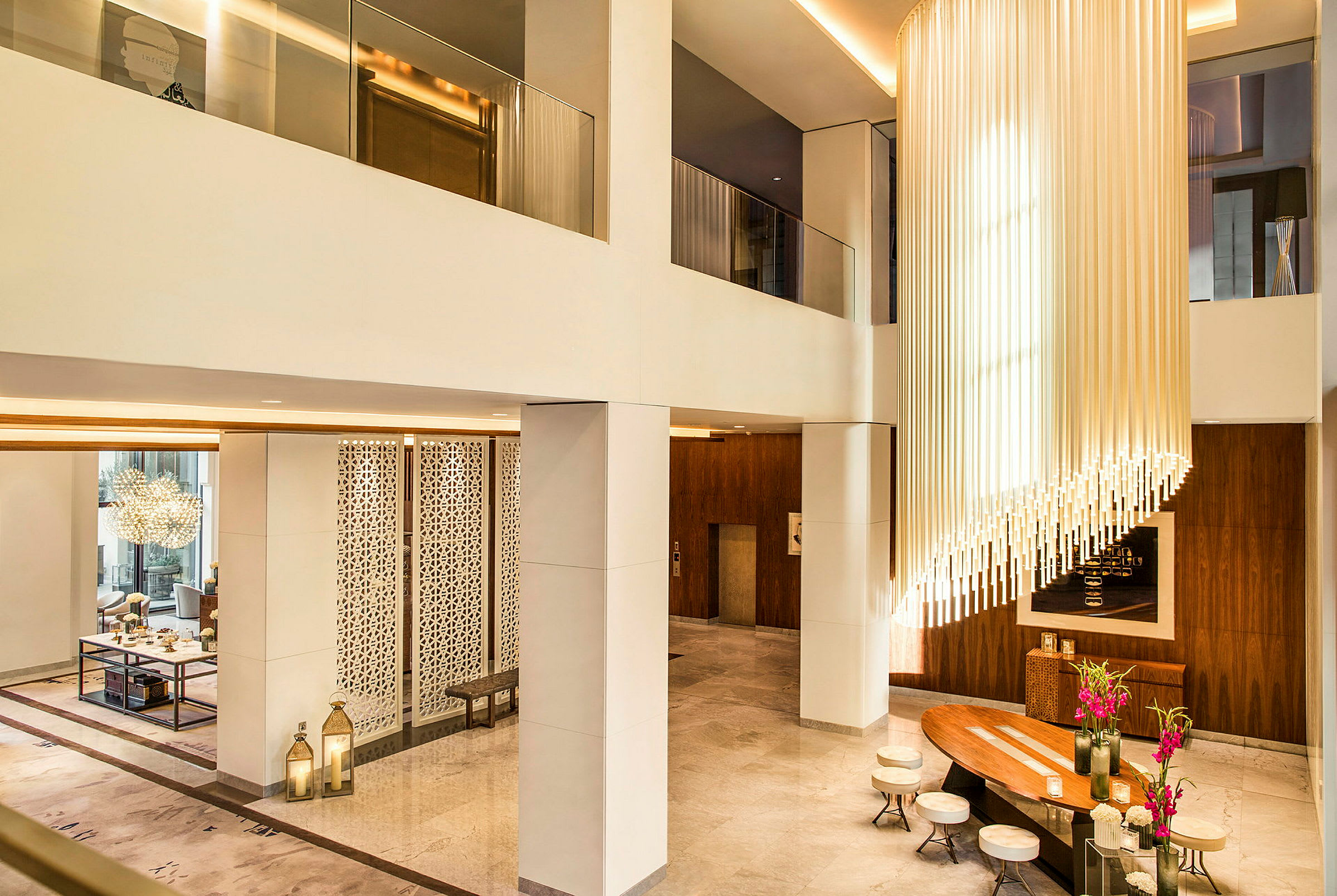 Ett elegant hotellobby med hög takhöjd, modern inredning och en ljuskrona av vertikala rör. Det finns sittplatser kring ett träbord med blommor. (AI-genererad text)