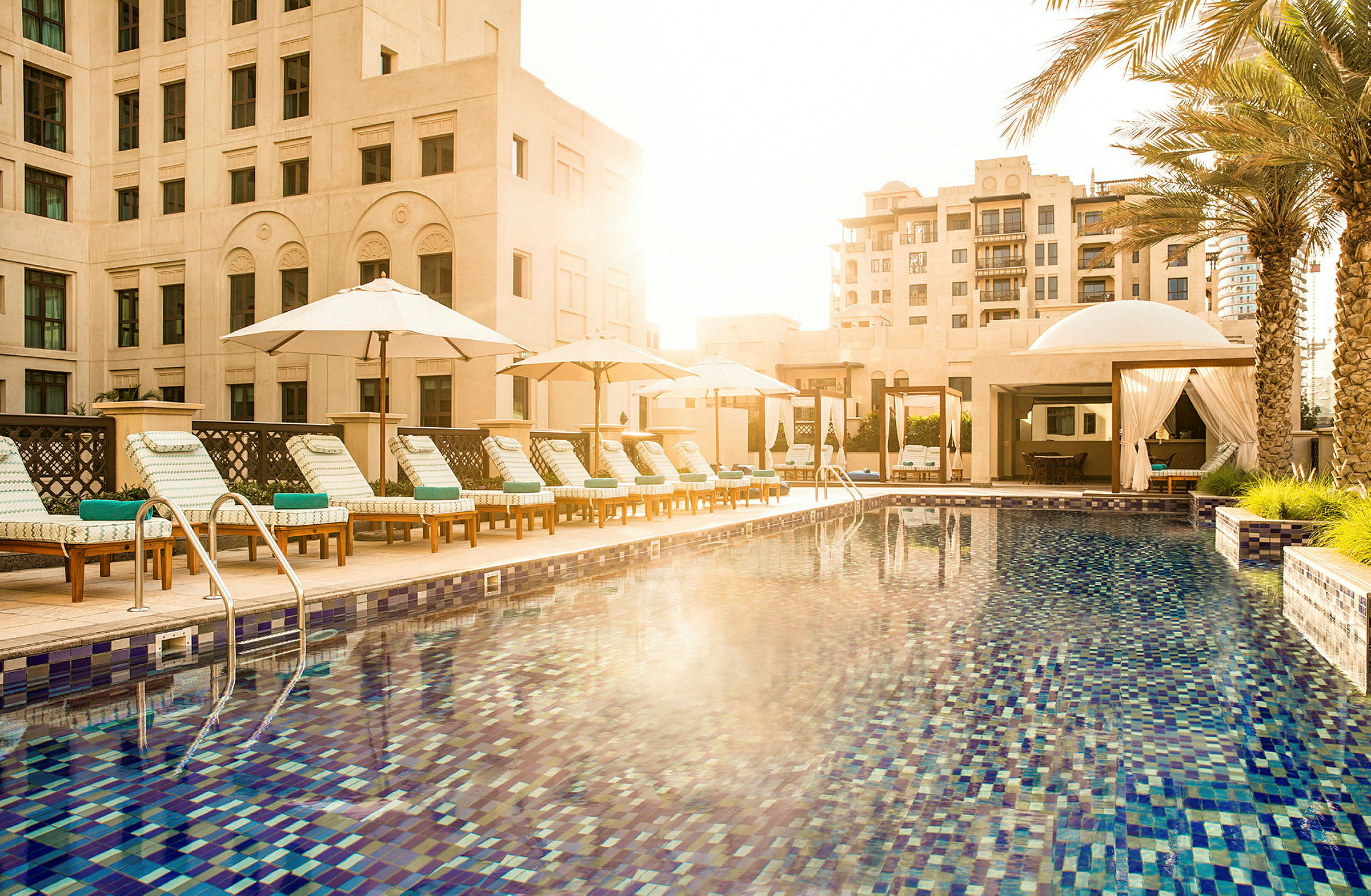 En hotellpool med solstolar och parasoller, omgiven av byggnader och palmer. (AI-genererad text)