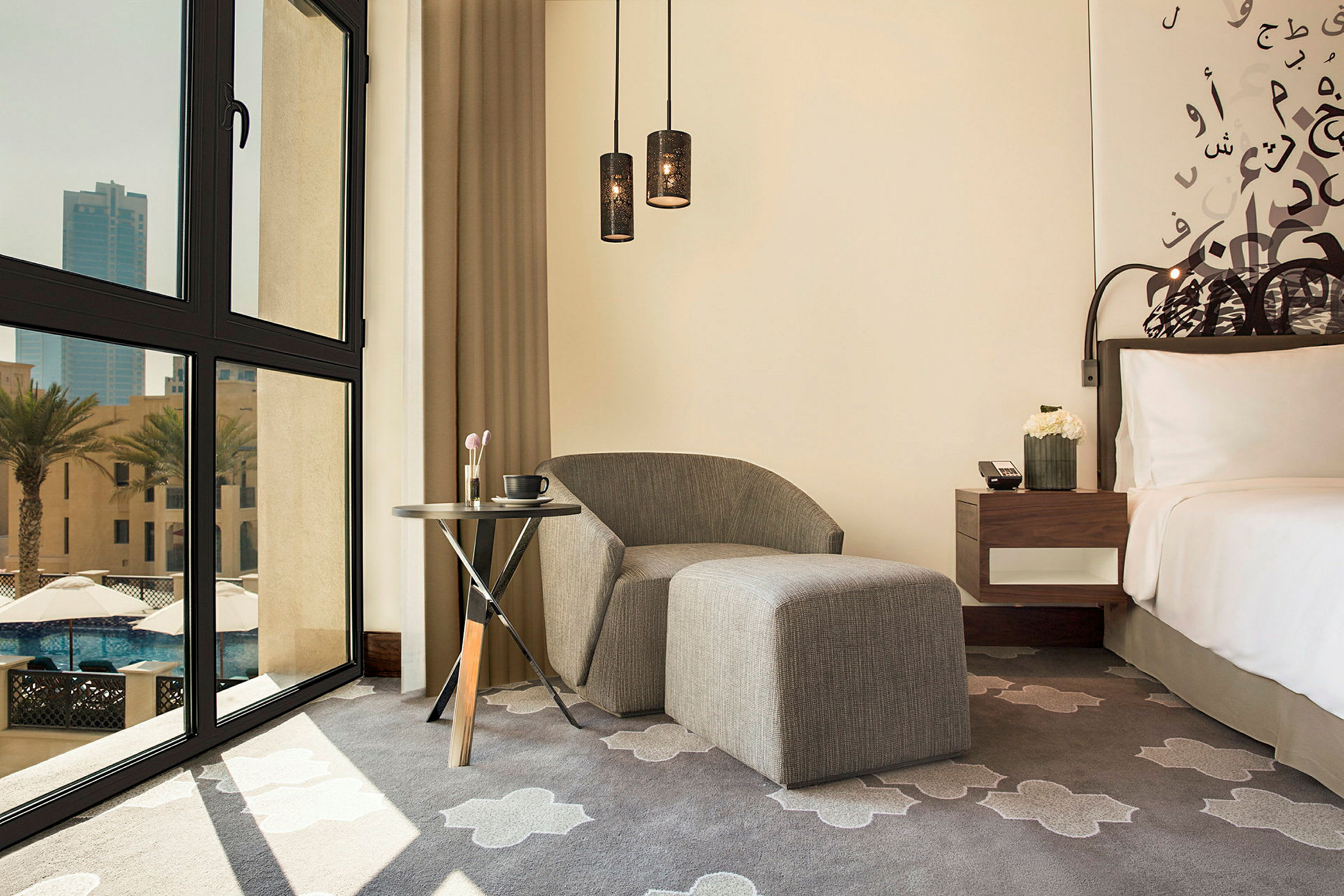Ett hotellrum med modern inredning. Bilden visar en grå stol med fotpall, ett litet bord med en kaffekopp och sminkborstar, samt en säng med vita sängkläder. Ett stort fönster med utsikt över palmträd och byggnader är också synligt. (AI-genererad text)