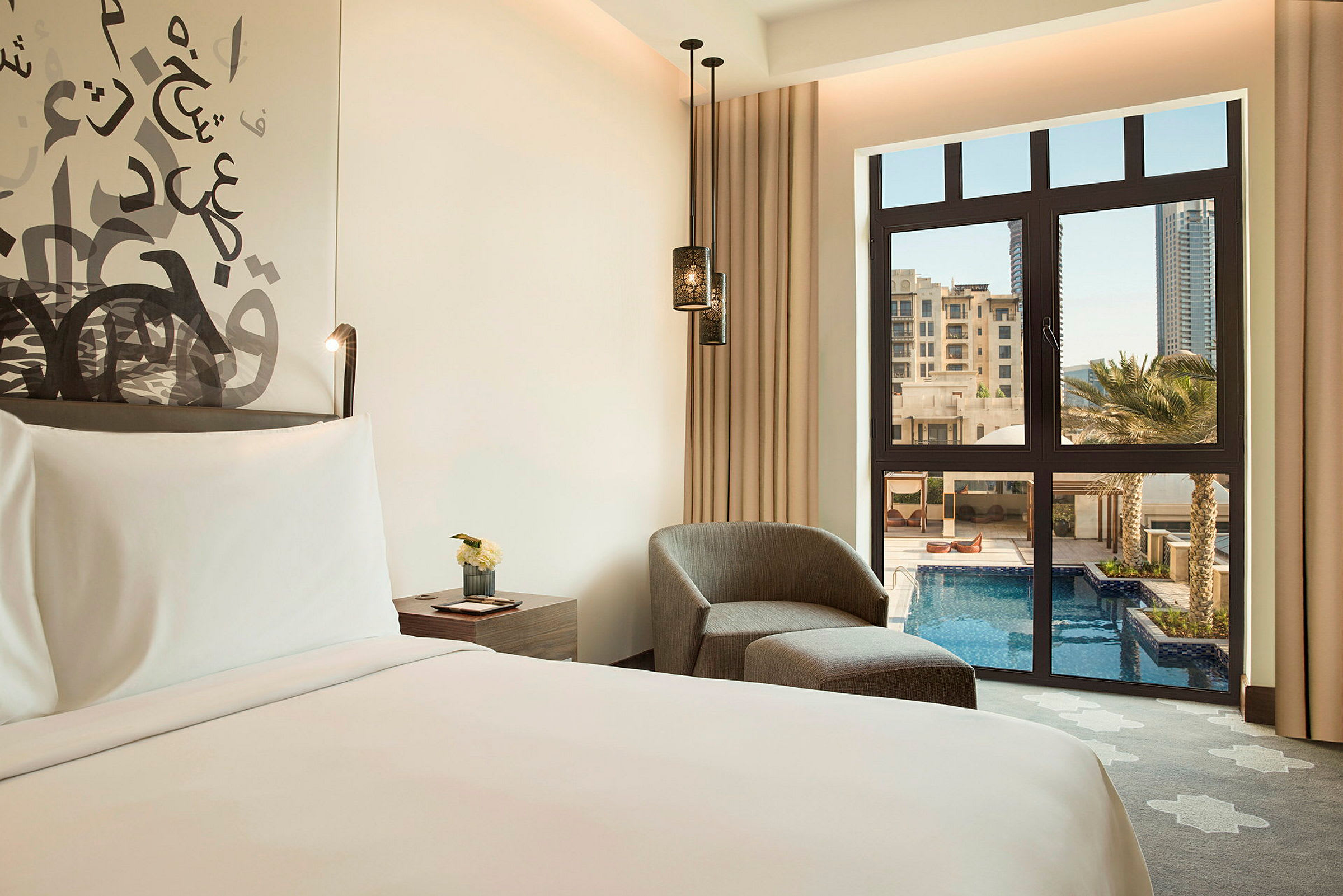 Bild av ett elegant hotellrum med en säng, en fåtölj och bord. Fönstret visar utsikt över en pool och byggnader. (AI-genererad text)