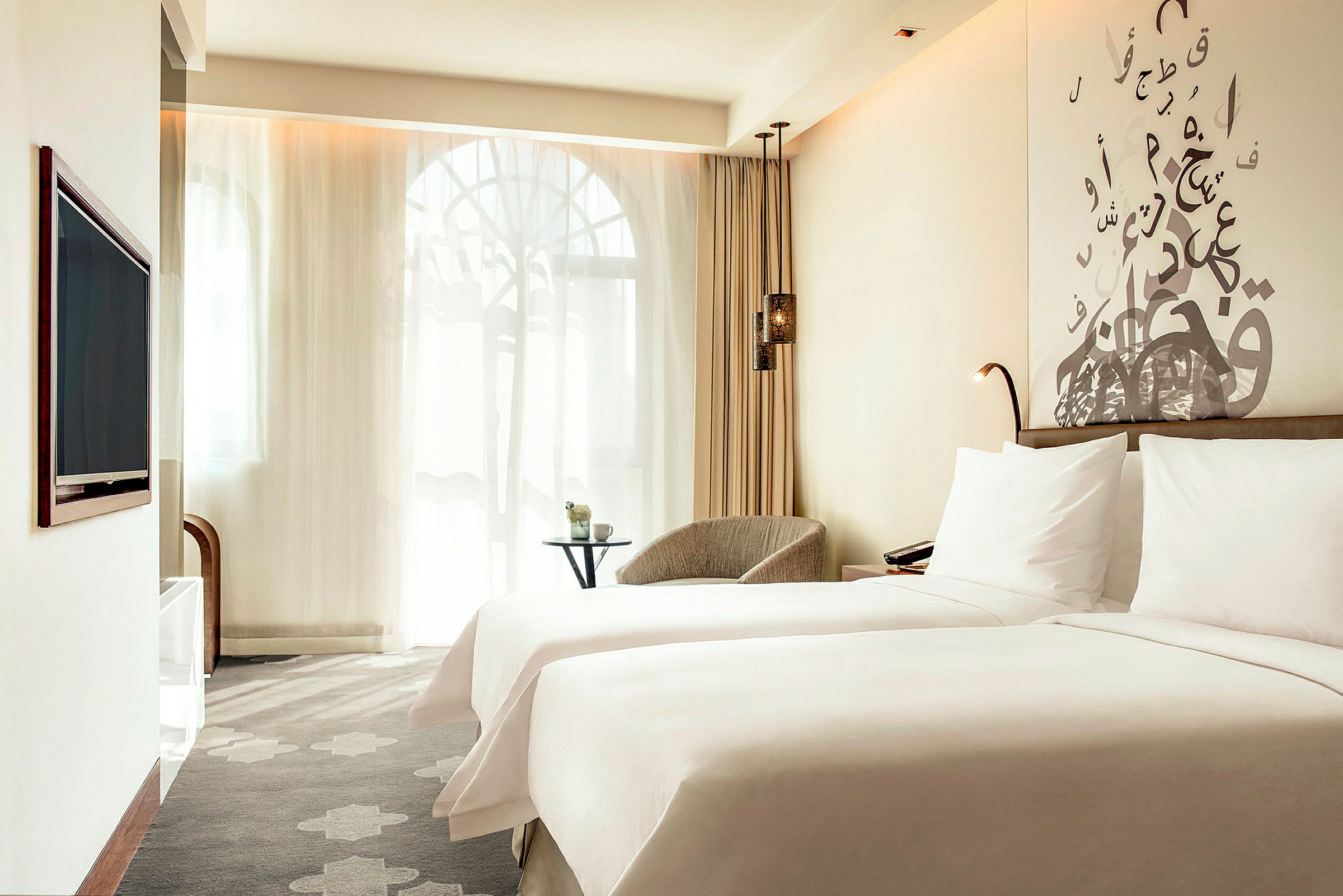 Ett hotellrum med två sängar som är bäddade med vita lakan. En sittplats med ett litet bord och fönster med tunna gardiner syns i bakgrunden. (AI-genererad text)