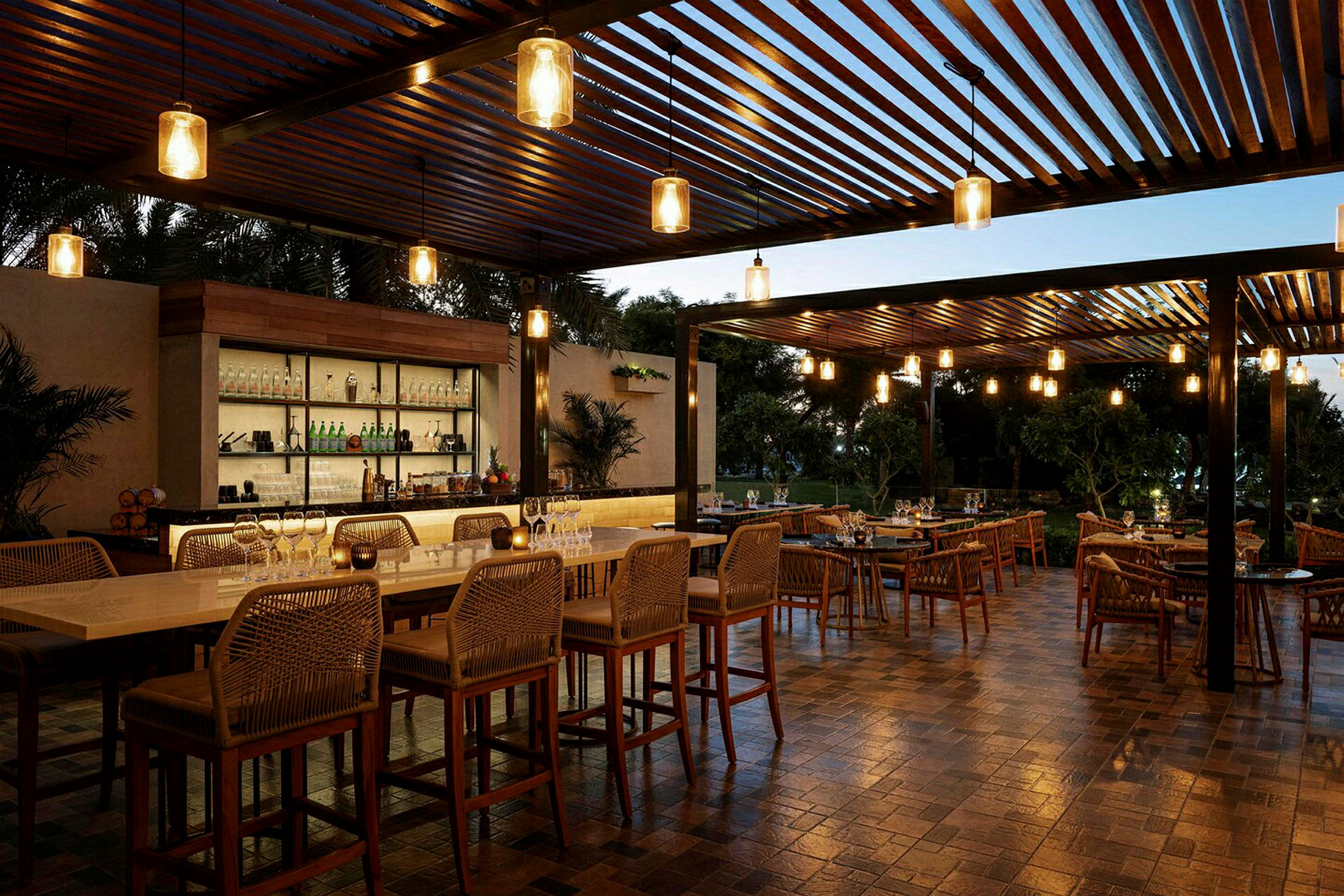 En utomhusrestaurang med bord och stolar samt en bar under en träpergola. Lampor hänger från taket och skapar en varm stämning. (AI-genererad text)