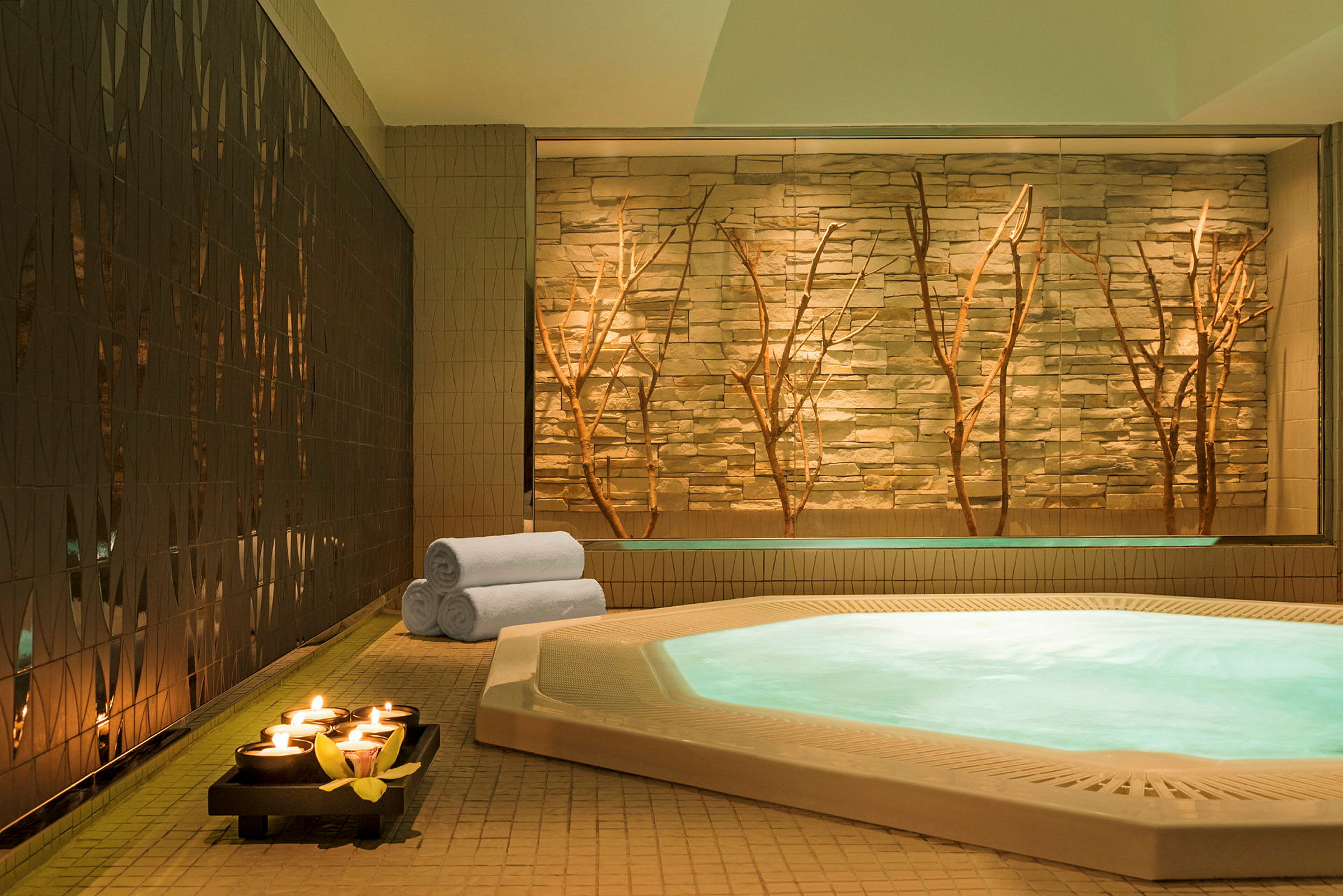 Ett spaområde med en upplyst jacuzzi, hoprullade vita handdukar och tända ljus på ett fat framför en dekorativ vägg med grenar. (AI-genererad text)