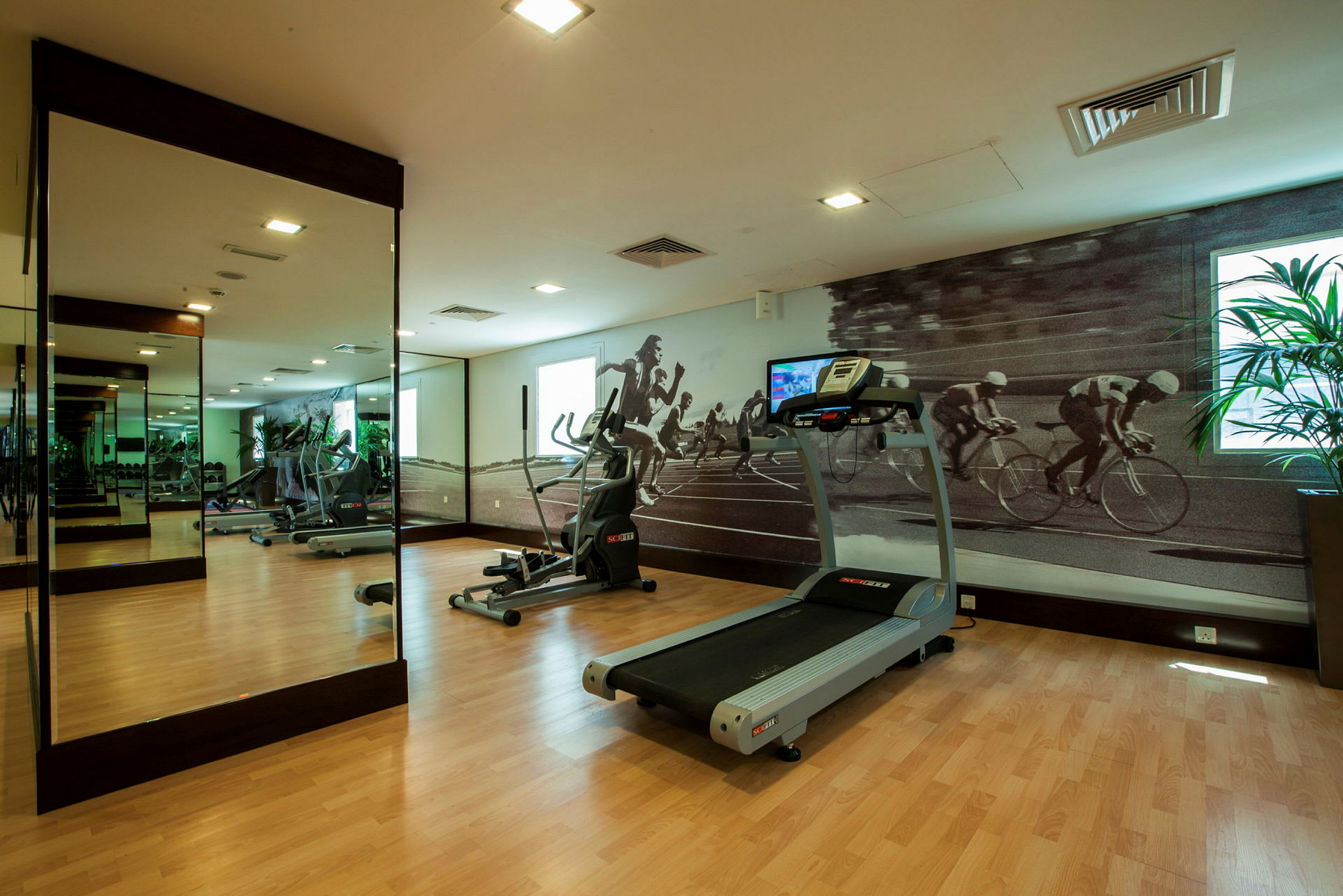 Ett gym med en löpband, en crosstrainer och en vägg med spegel och motiv av cyklister och löpare. (AI-genererad text)