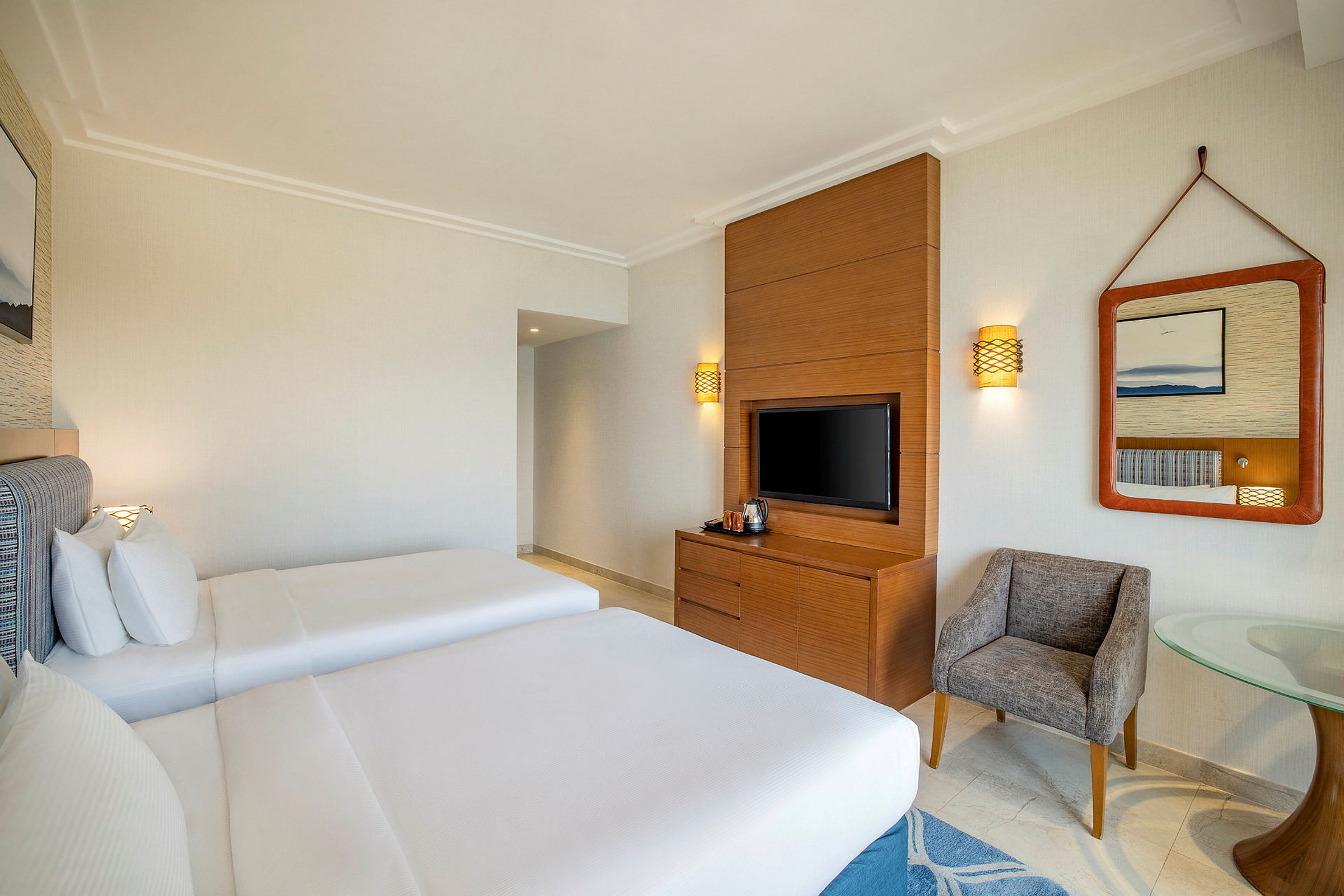 Ett hotellrum med två sängar, en TV på en träpanel, en spegel och en liten sittgrupp. (AI-genererad text)