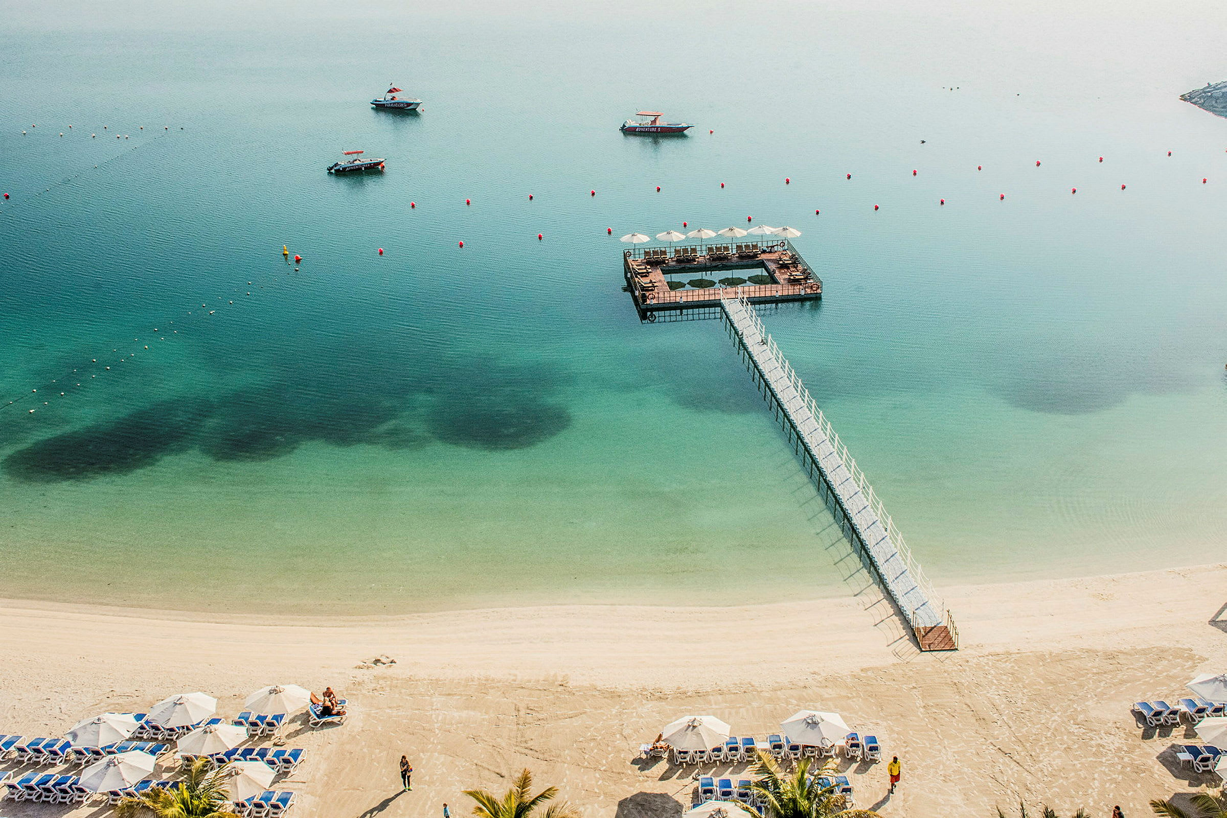 Strand med parasoller, solstolar och en brygga ut till en flytande plattform på havet. (AI-genererad text)
