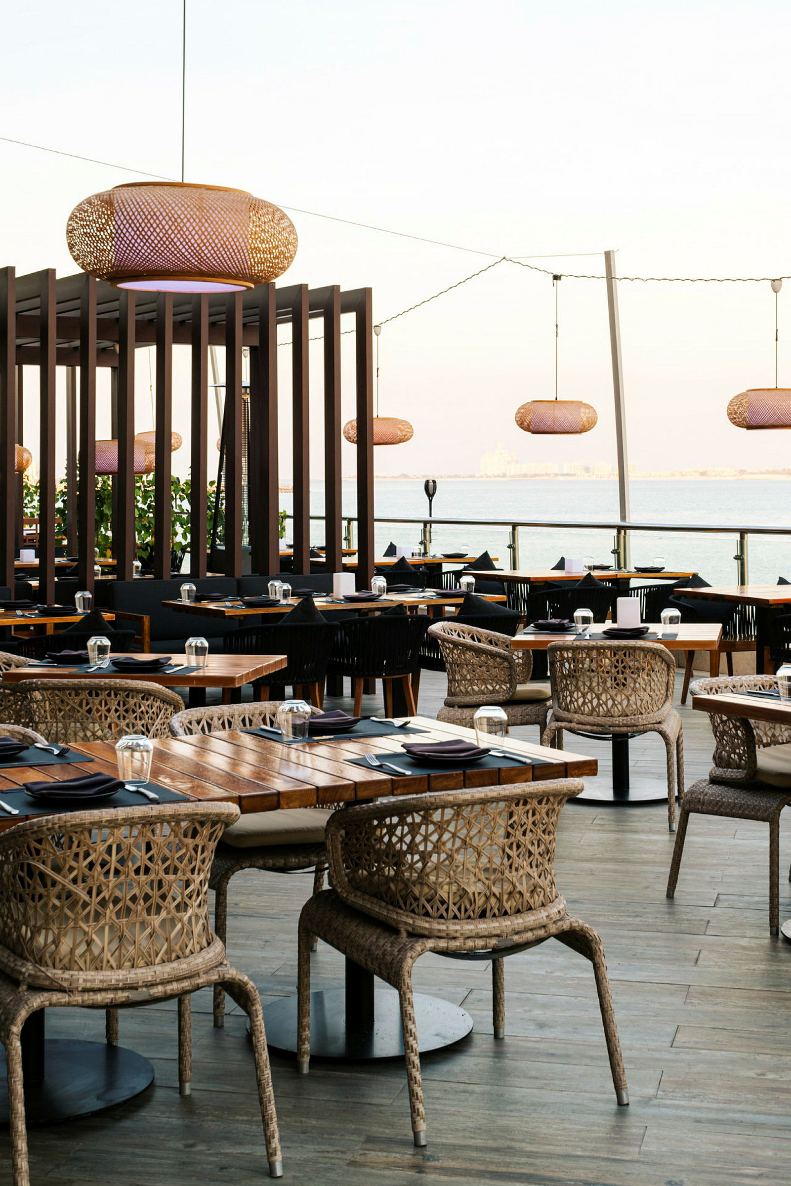 En utomhusrestaurang med trämöbler, rottingstolar och bord förberedda med svarta servetter och glas. Framför havet. (AI-genererad text)