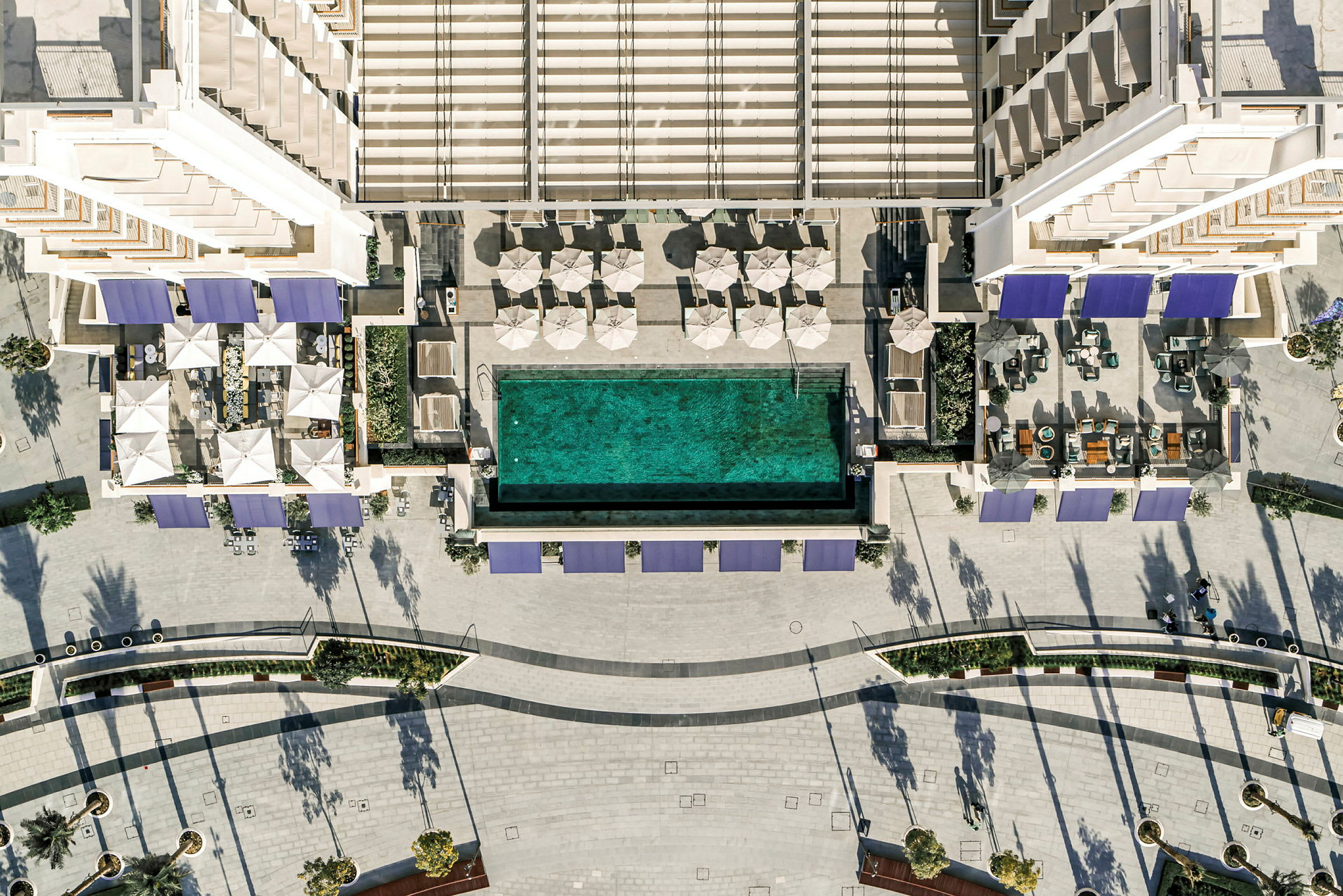 Topphotografisk vy av en modern byggnad med en avlång swimmingpool omgiven av parasoller och loungemöbler samt ett öppet stenlagt område. (AI-genererad text)
