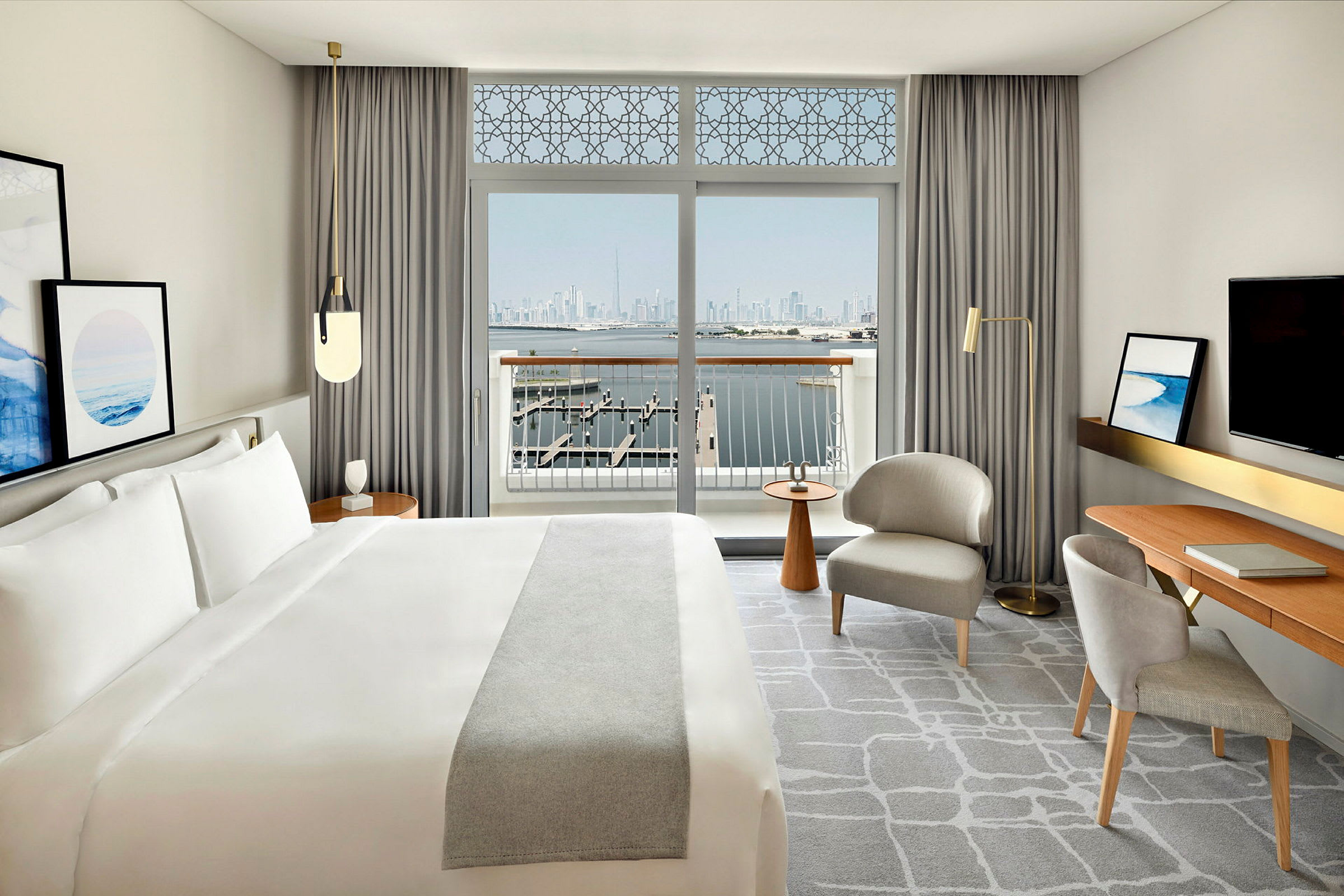 Hotellrum med en stor säng, skrivbord, sittplatser och en balkong med utsikt över vatten och en stadshorisont. (AI-genererad text)