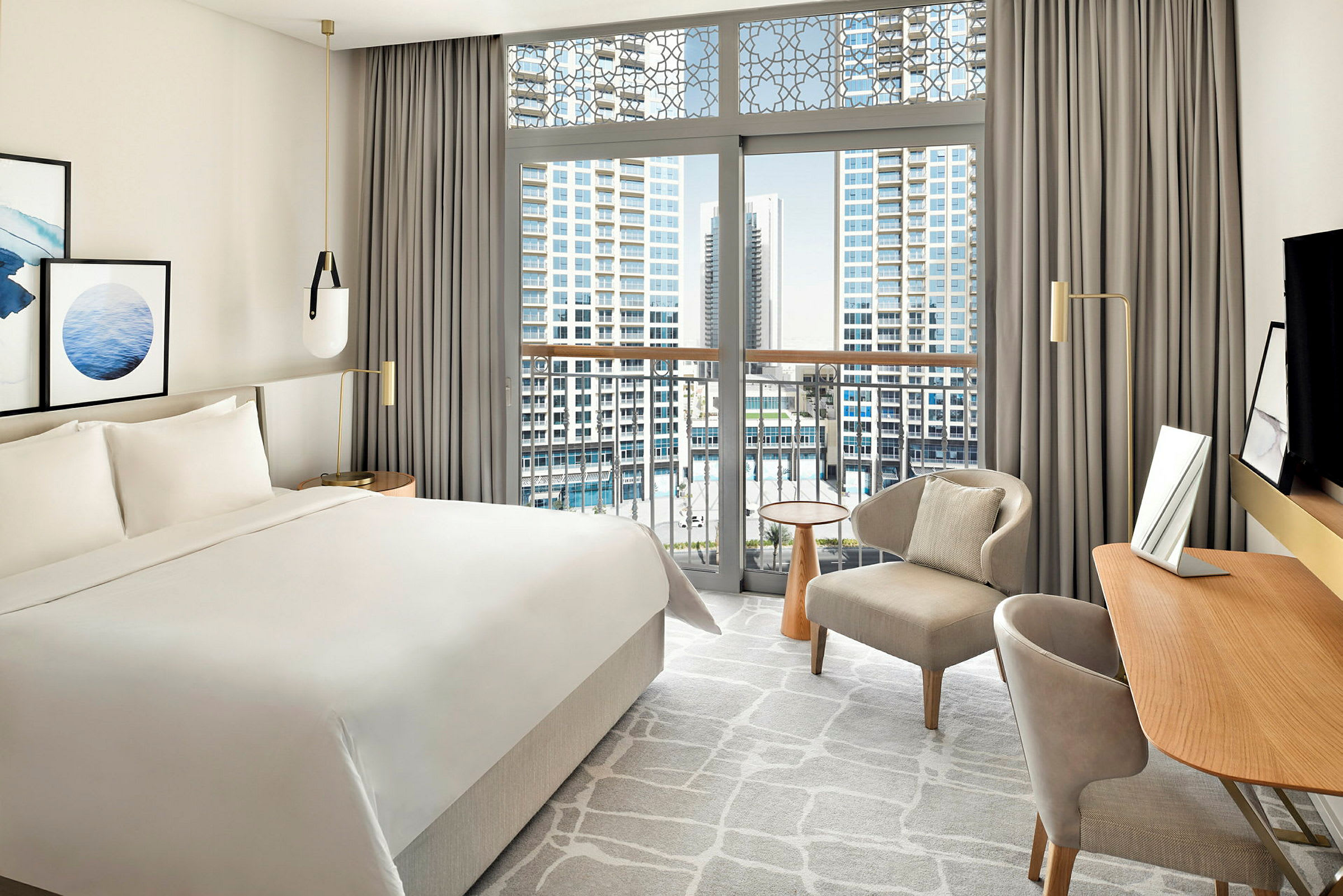 Ett modernt hotellrum med en stor säng, skrivbord, stol och ett stort fönster som erbjuder utsikt över en stad med höga byggnader. (AI-genererad text)