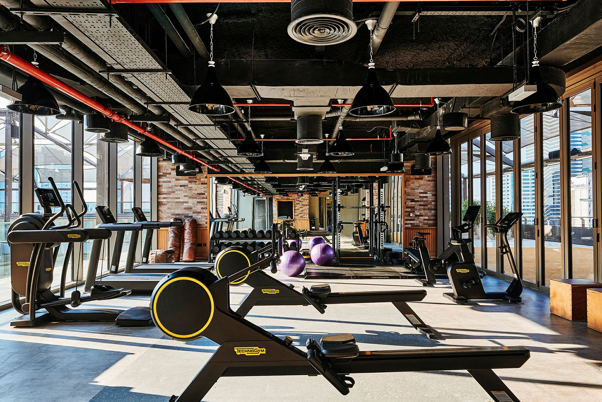 Bild på ett gym med träningsredskap som löpband, roddmaskiner och hantlar, samt stora fönster. (AI-genererad text)