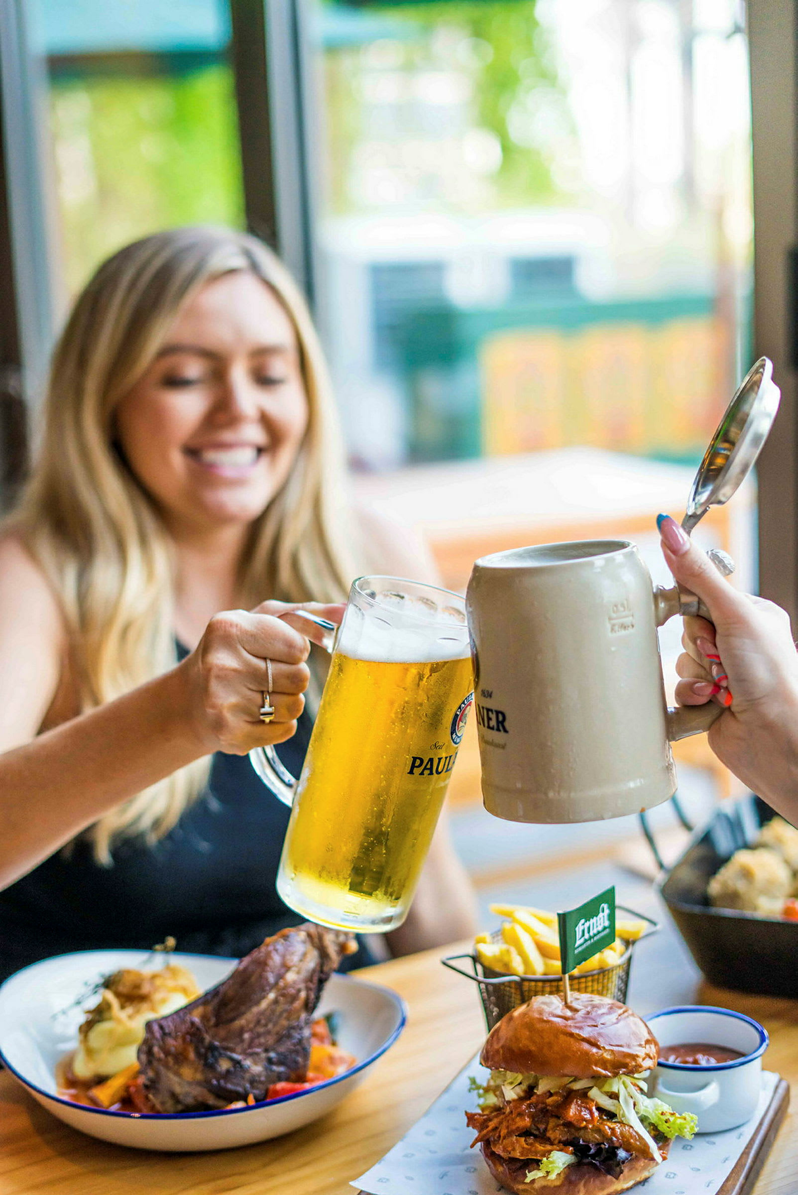 Två fyllda ölglas skålas, och på bordet finns tallrikar med mat inklusive hamburgare, pommes frites och köttbit. (AI-genererad text)