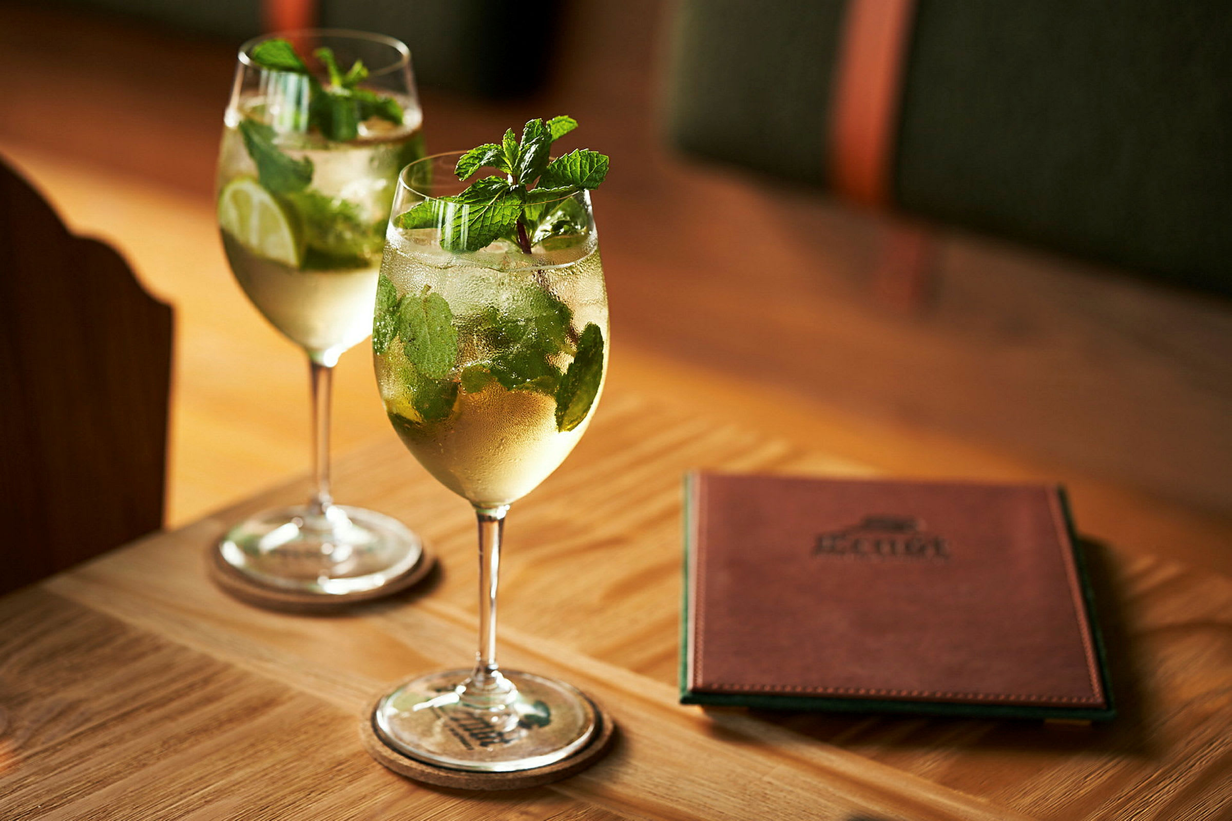 Två glas med drinkar innehållande mint och lime står på ett bord bredvid ett menyhäfte. (AI-genererad text)