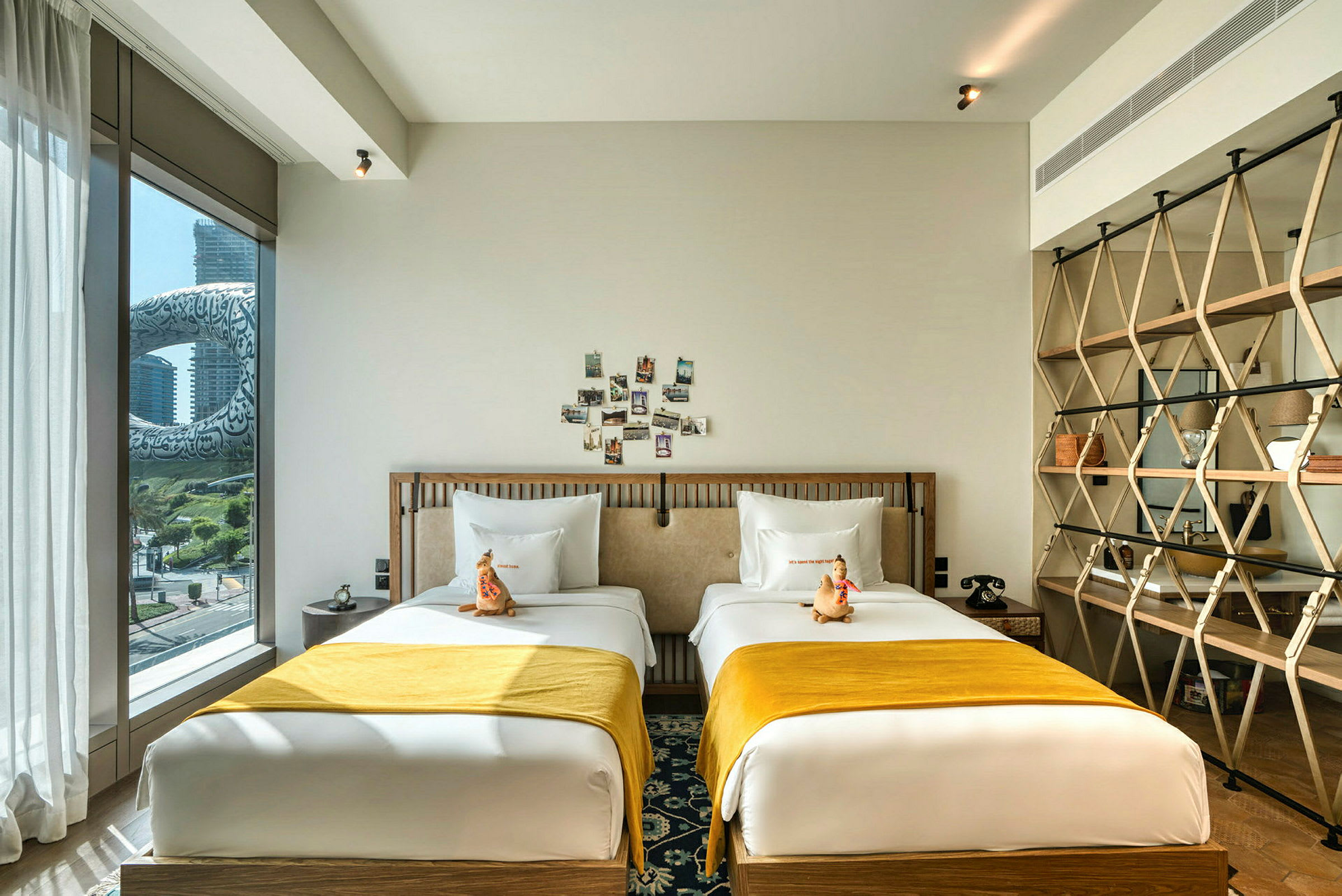 Ett hotellrum med två enkelsängar med gula överkast, dekorativa kuddar och ett fönster som visar en utsikt över en modern byggnad utanför. (AI-genererad text)