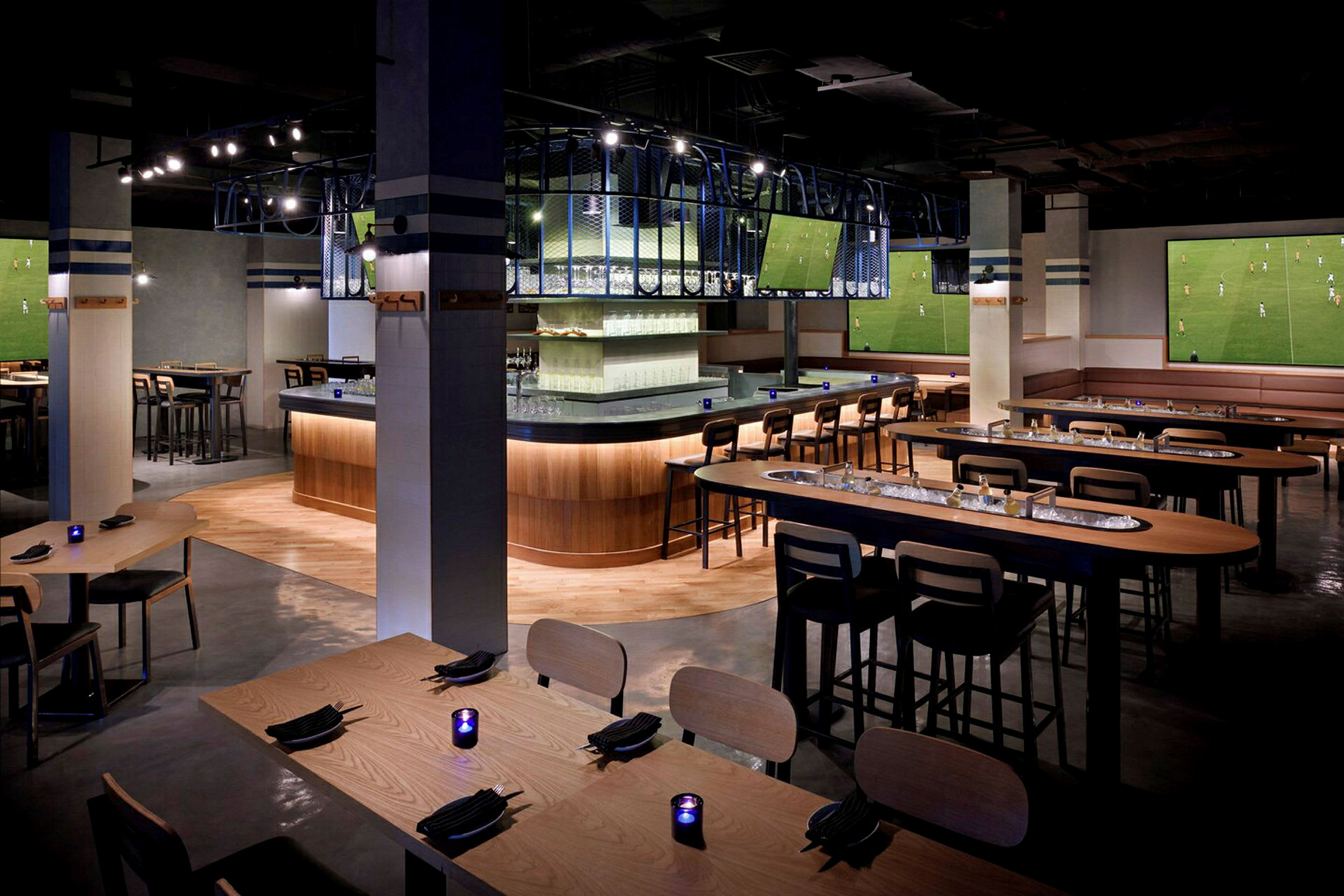 Interiör av en modern bar med sittgrupper, flera bord och stolar, samt stora skärmar på väggarna. (AI-genererad text)