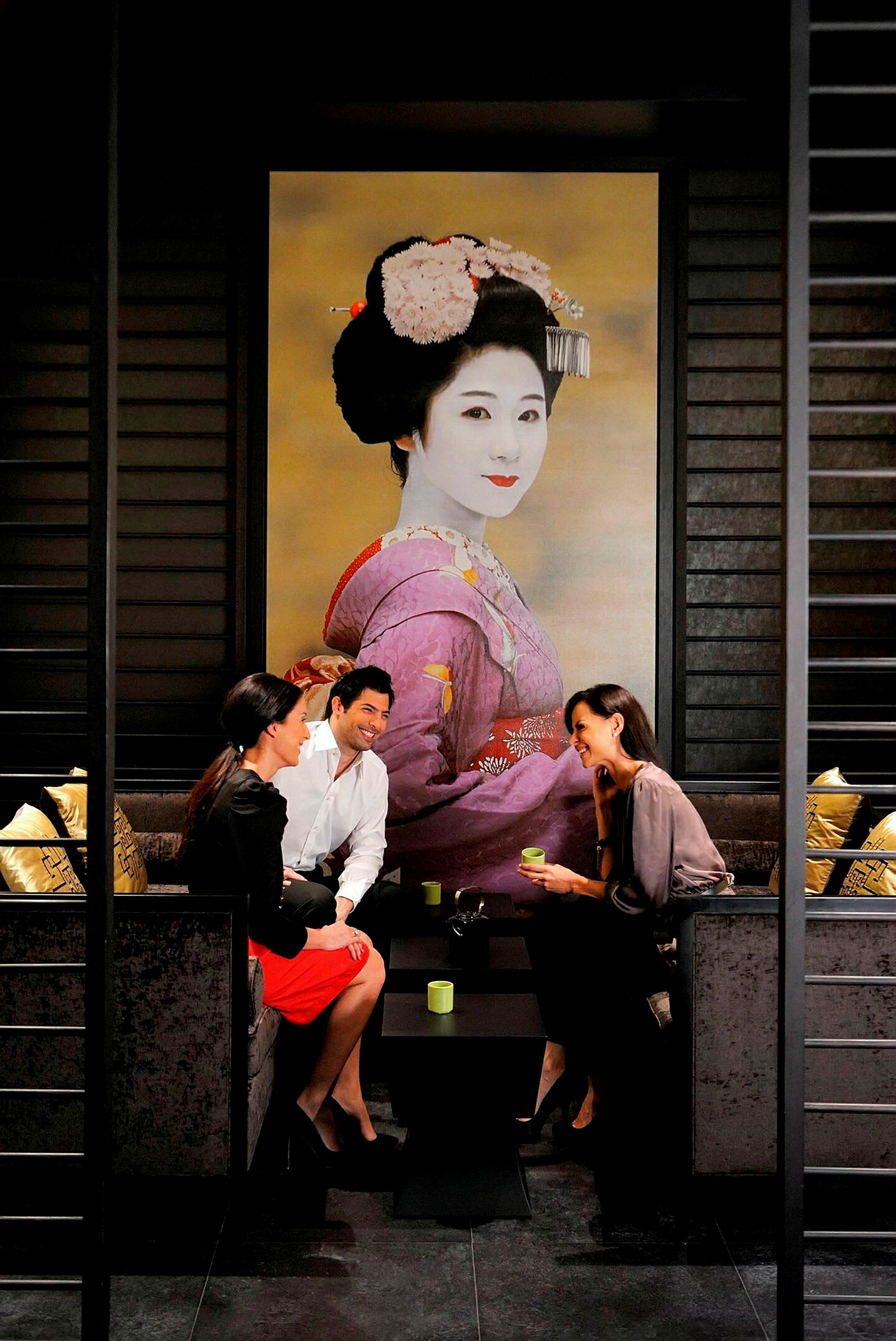 Tre personer sitter vid ett bord i en elegant miljö med en stor bild av en geisha i bakgrunden. (AI-genererad text)