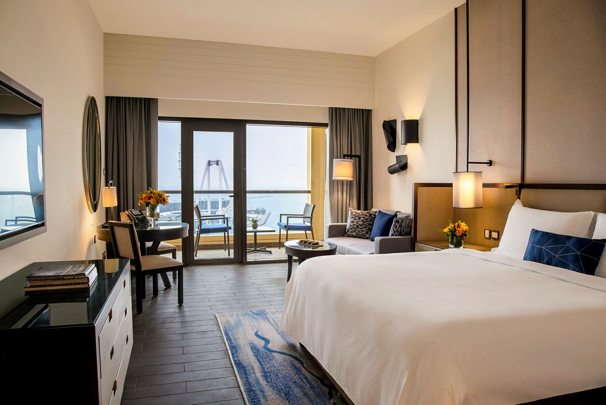 Ett hotellrum med en stor säng, sittgrupp och en matplats. Stora fönster leder ut till en balkong med utsikt över havet och ett stort pariserhjul. (AI-genererad text)