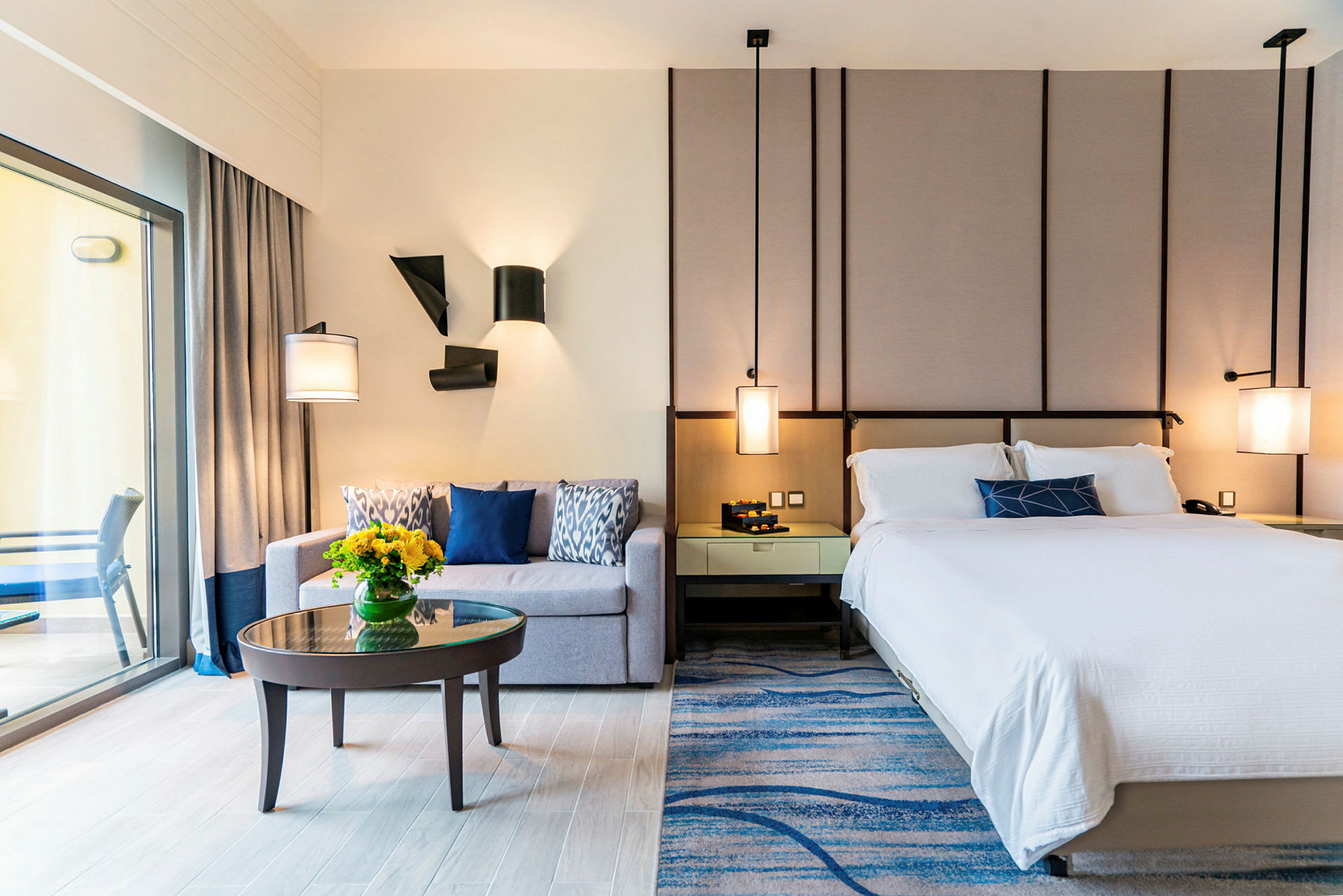 Ett modernt hotellrum med en säng, en soffa, ett litet soffbord med en vas med blommor och en balkong som syns genom en glasdörr. (AI-genererad text)