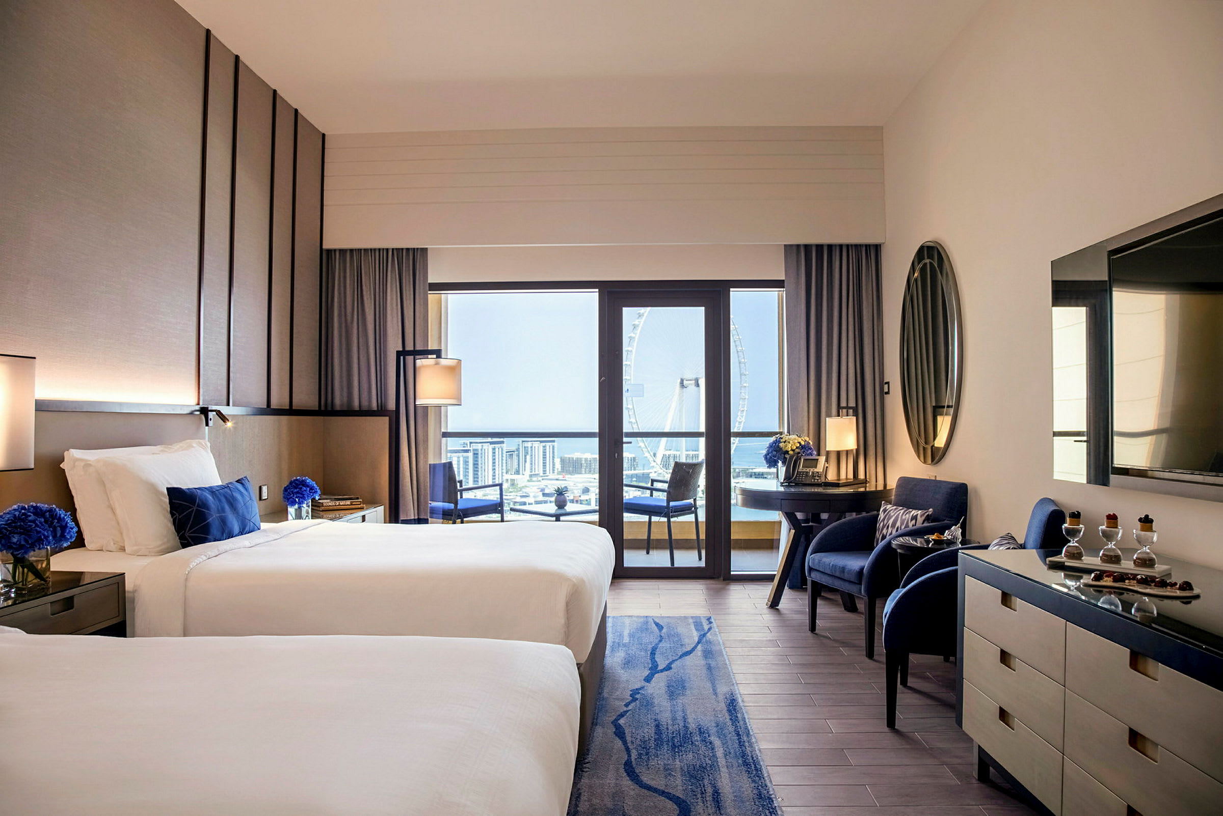 Ett hotellrum med två enkelsängar, en skrivbordsdel, två stolar vid sidan samt en balkong med utsikt över ett pariserhjul och en stadssilhuett. (AI-genererad text)