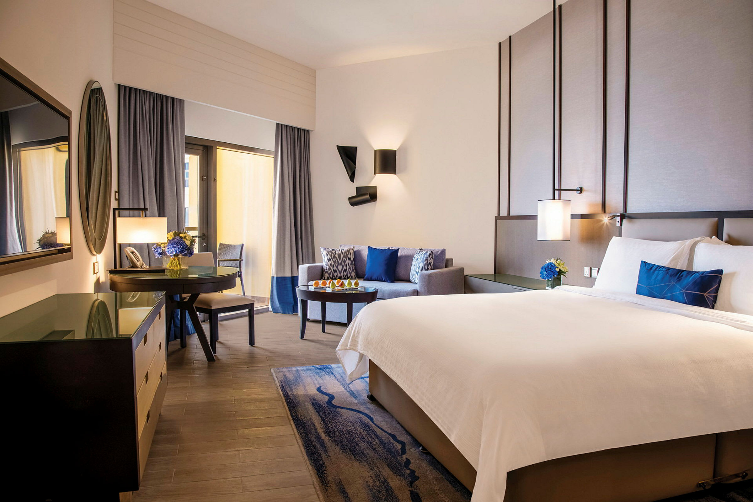 Hotellrum med en stor dubbelsäng, sittgrupp, skrivbord och en TV. Rummet är elegant inrett med neutrala färger och blå detaljer. (AI-genererad text)