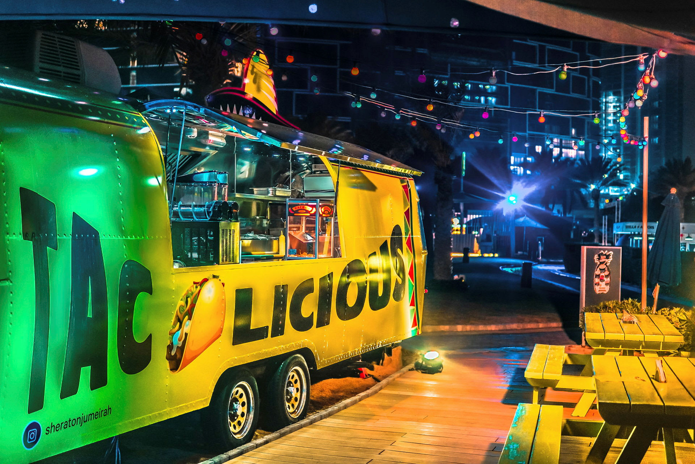 En gul matvagn med texten 'Tacolicious', nattmiljö med lysande dekorationer. (AI-genererad text)