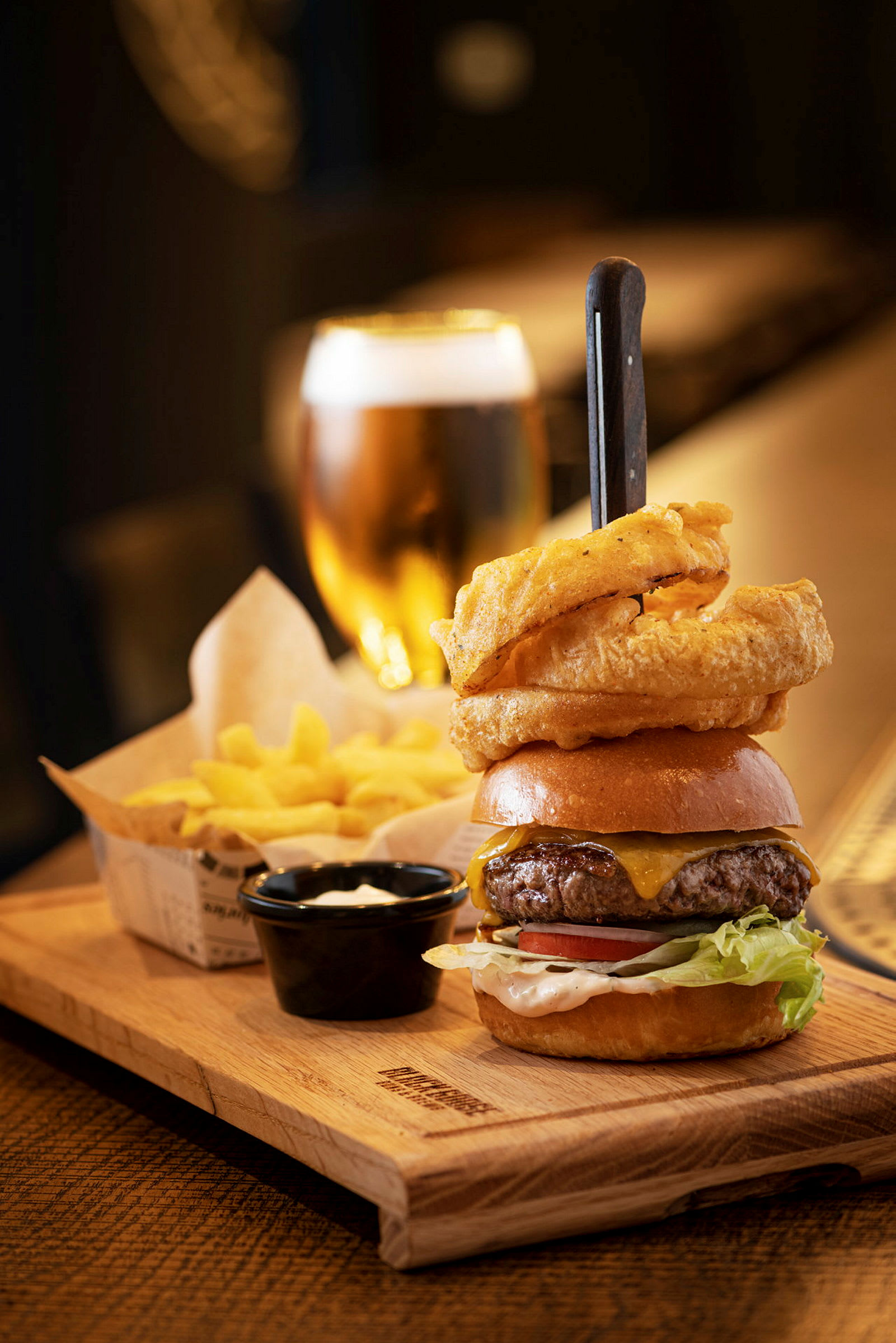En tallrik med en hamburgare, lökringar, pommes och en dippsås. En öl i glas syns i bakgrunden. (AI-genererad text)