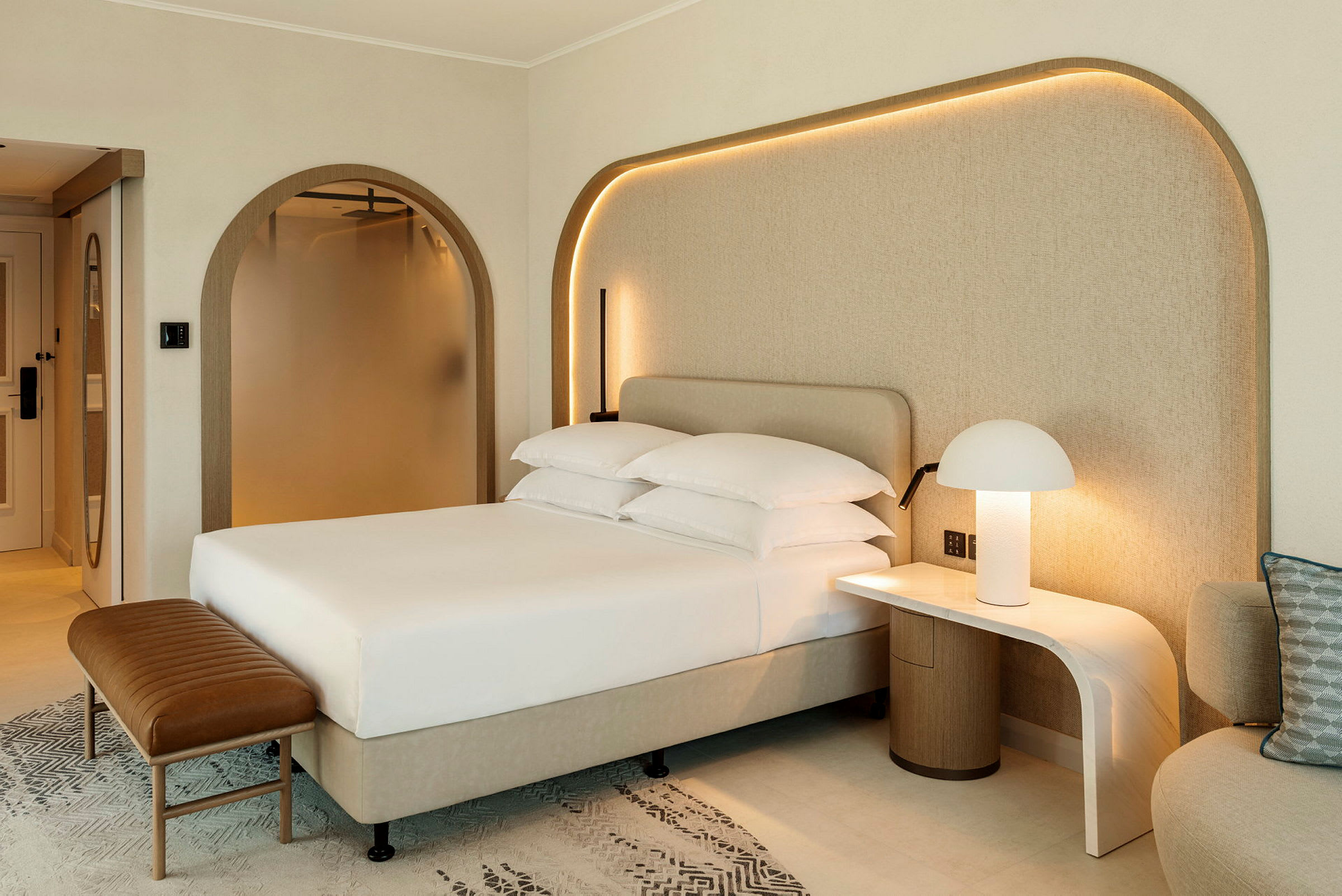 Bild på ett modernt hotellrum med en bäddad säng, nattduksbord med lampa och en sittmöbel. (AI-genererad text)
