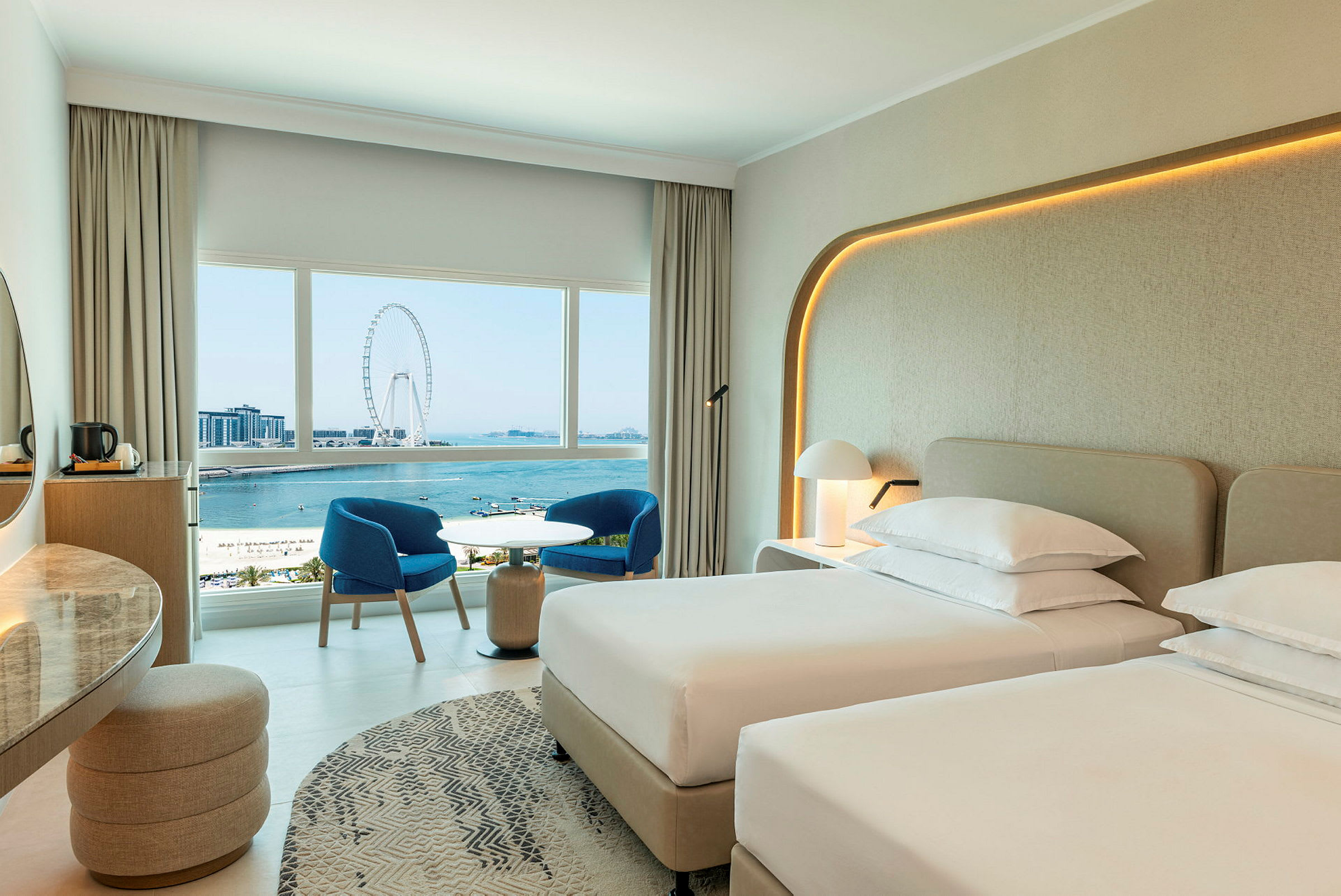 Hotellrum med två enkelsängar, en sittgrupp med två blå stolar och ett bord, samt utsikt över havet och ett stort pariserhjul. (AI-genererad text)