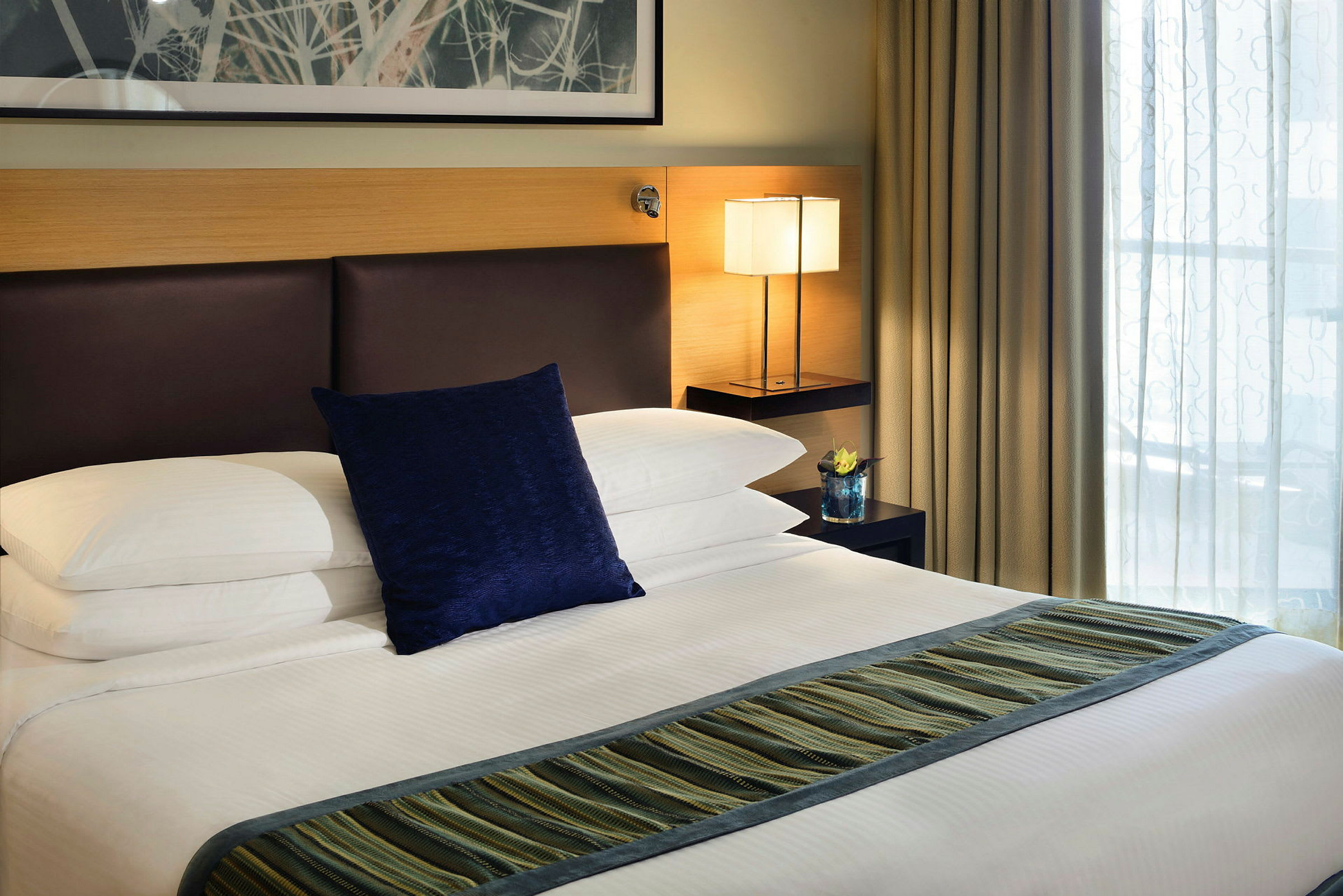 En hotellrumssäng med vita lakan, en blå kudde och en randig löpare. Ett nattduksbord med en lampa och en dekoration är placerad på sidan. (AI-genererad text)