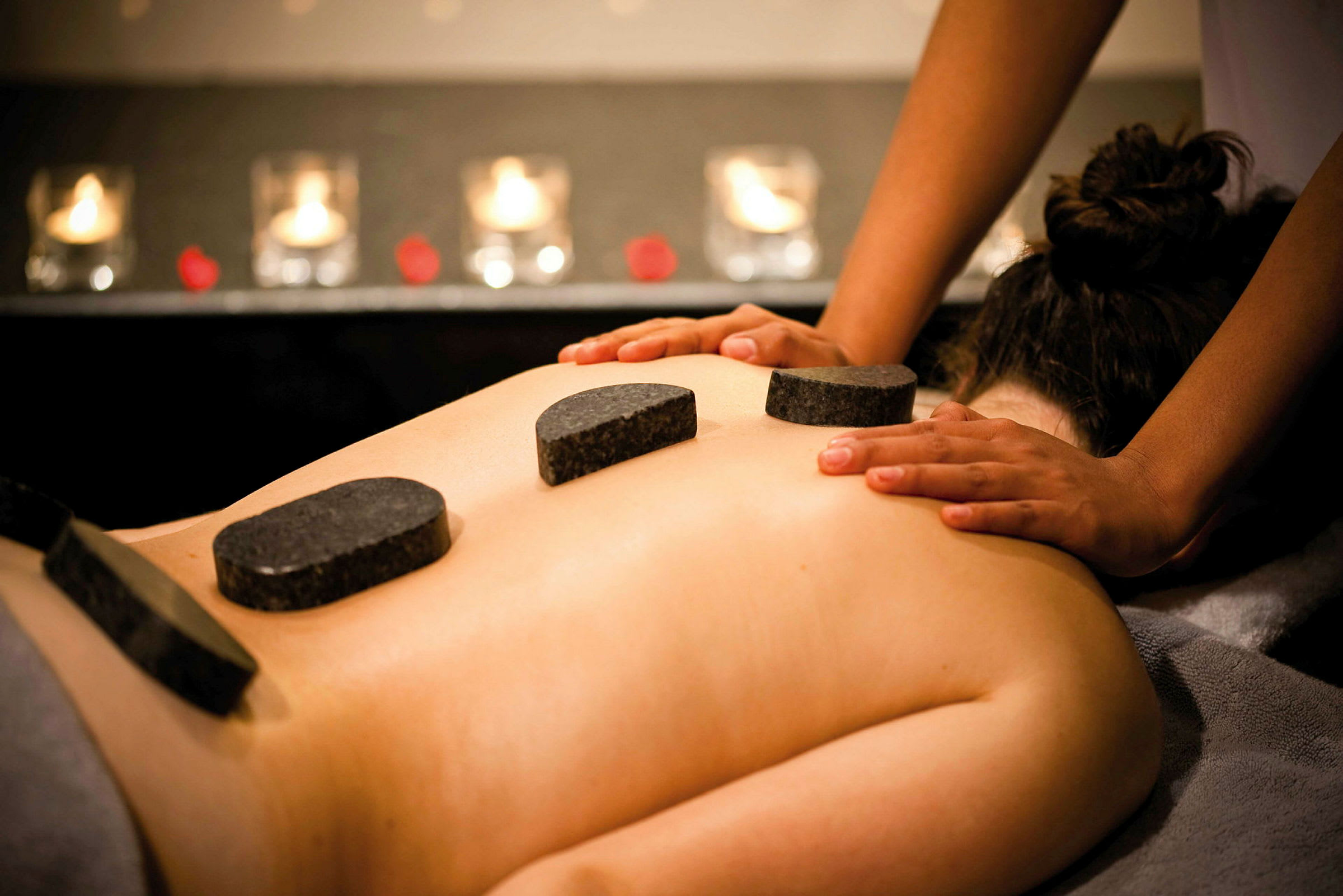 En person får hot stone-massage, med varma stenar placerade på ryggen samt händer som ger massage. (AI-genererad text)