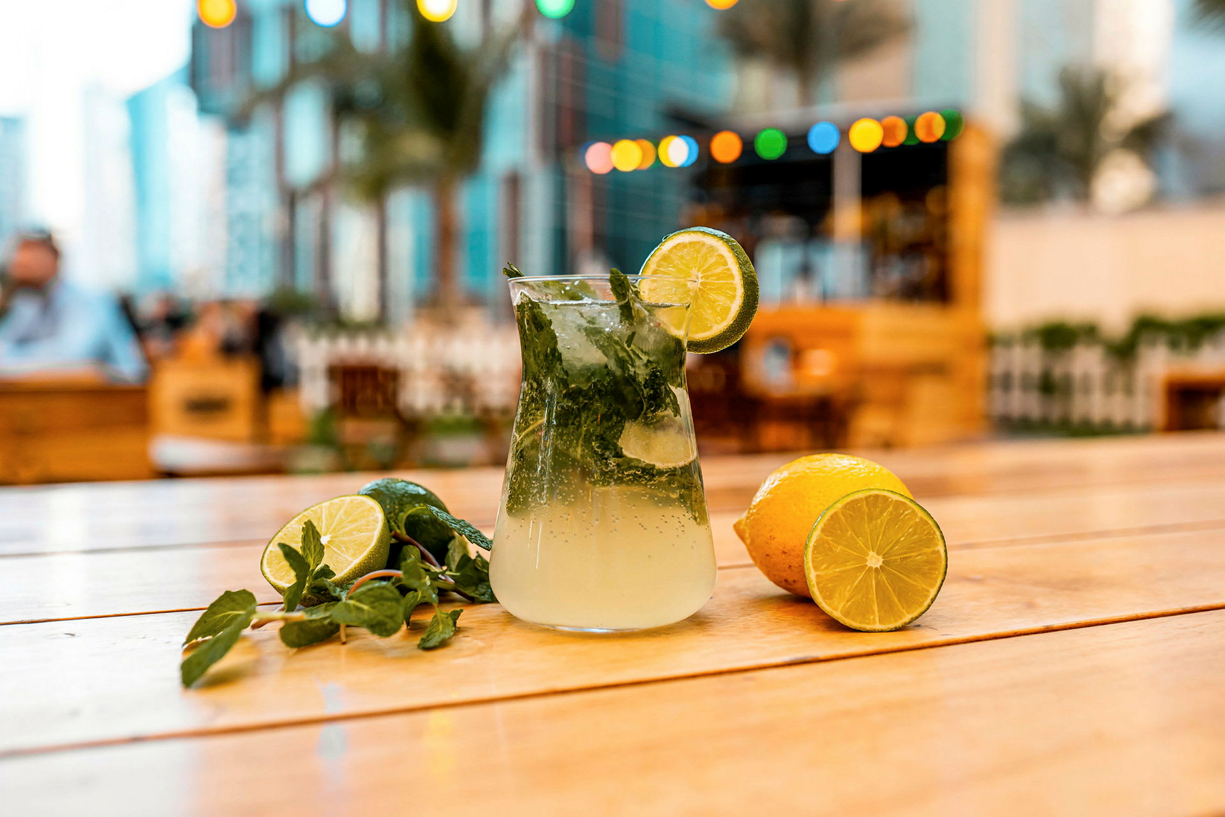 En glasbehållare med en uppfriskande citron- och mintdryck står på ett träbord, omgiven av färska citronskivor och mintblad. Bakgrundsmiljön är en utomhusområde med dekorationer. (AI-genererad text)