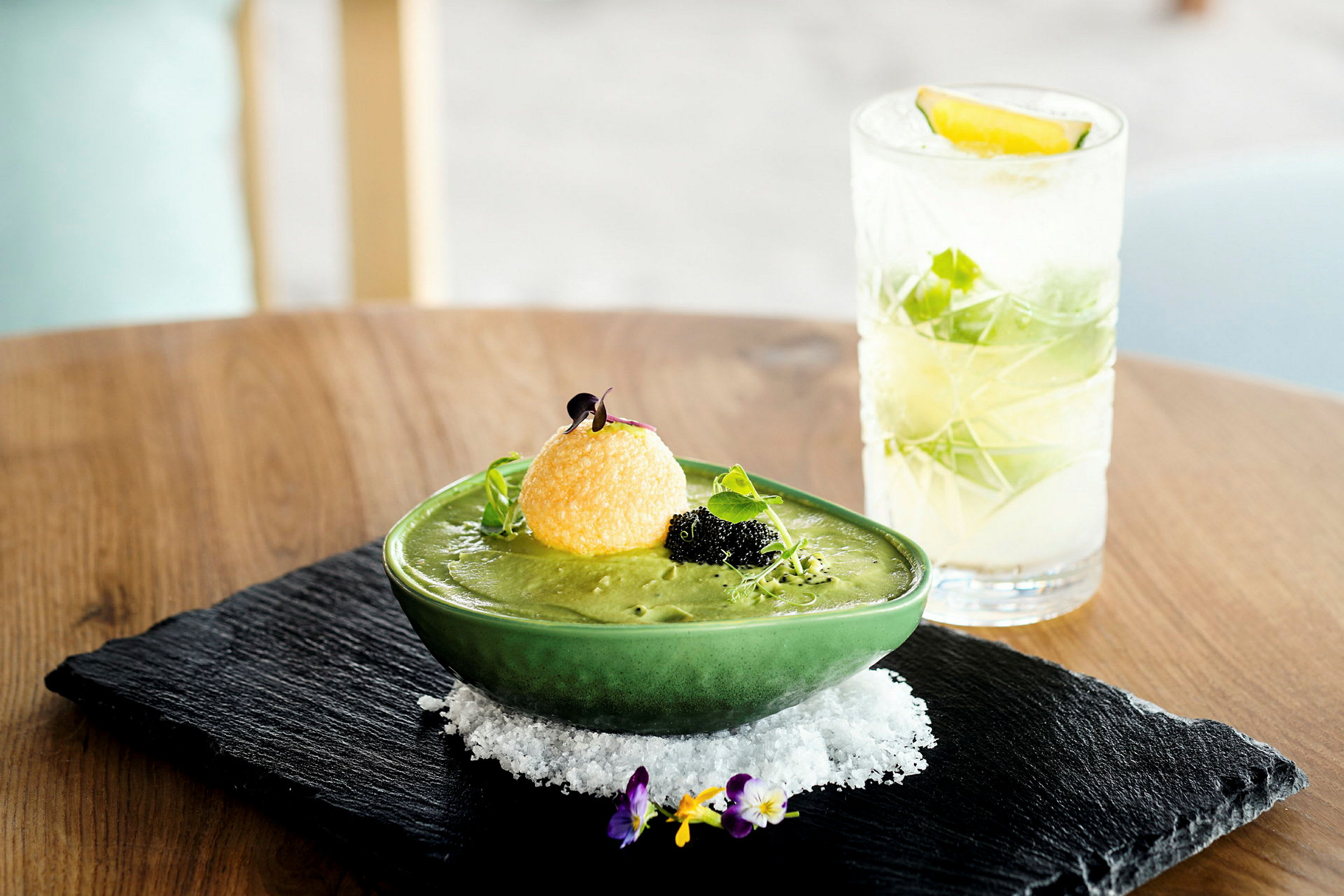 En grön soppa i en skål med garnityr bredvid en cocktail på ett bord. (AI-genererad text)