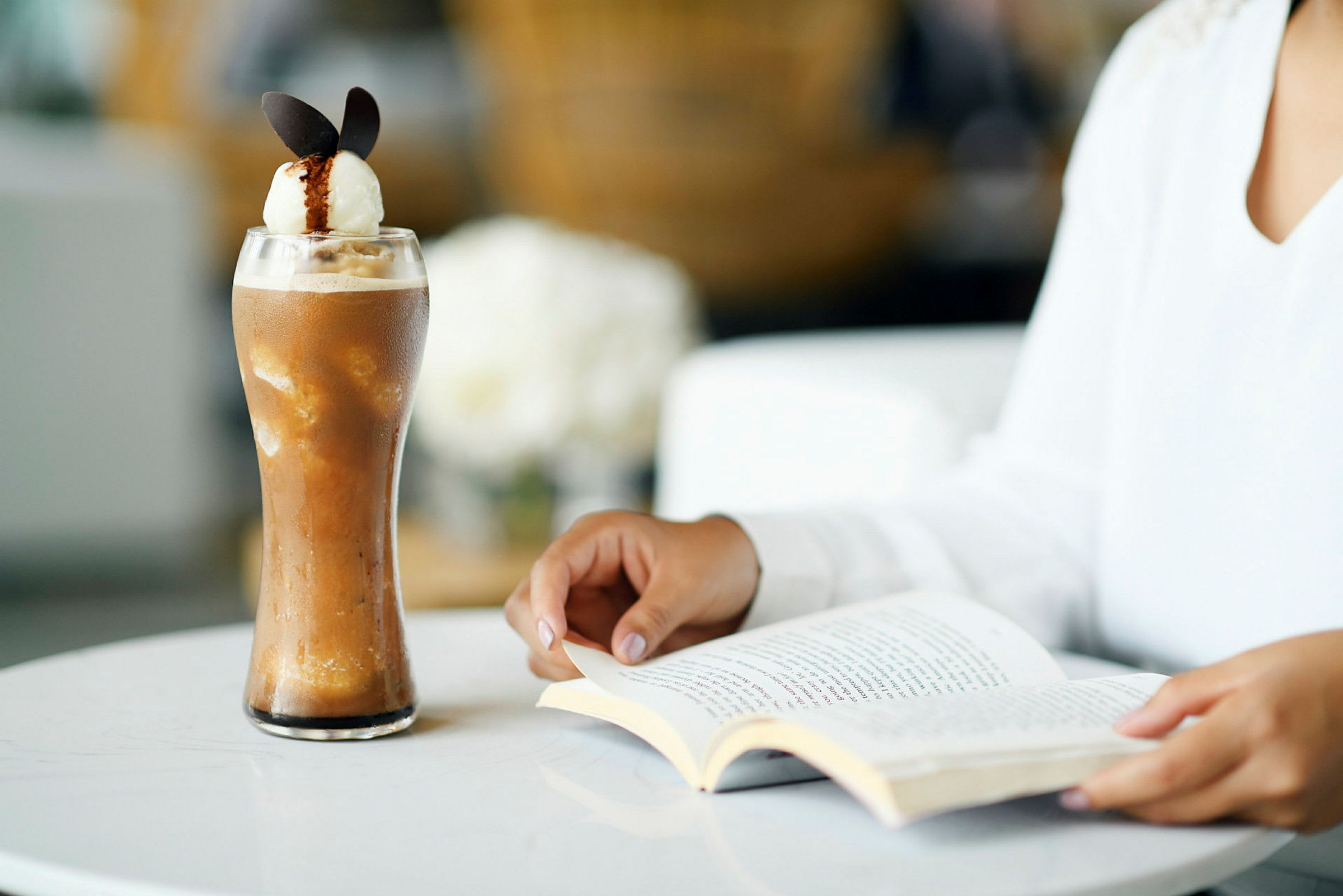 Ett glas med kall kaffedryck och en person som läser en bok vid ett bord. (AI-genererad text)