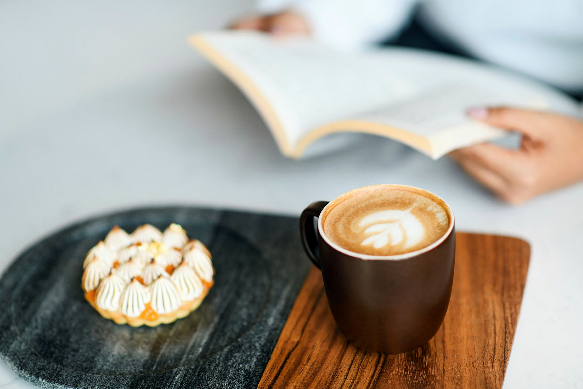 En kopp kaffe och en liten bakelse ligger på en träbricka. En person håller en öppen bok i bakgrunden. (AI-genererad text)