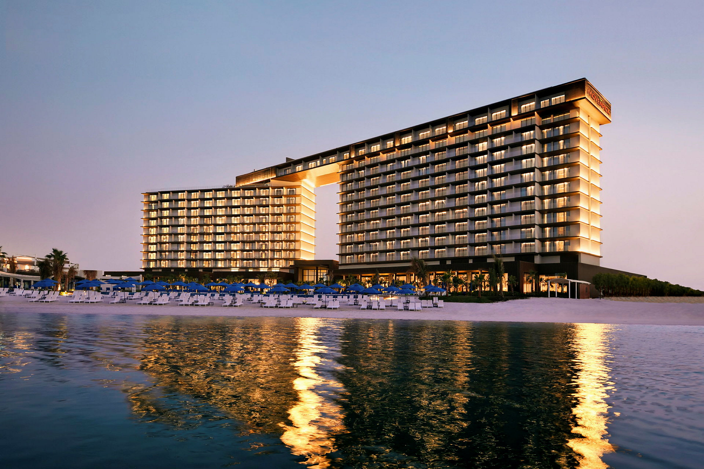 Byggnaden är ett modernt hotellkomplex vid en strand, upplyst och speglar sig i vattnet. (AI-genererad text)