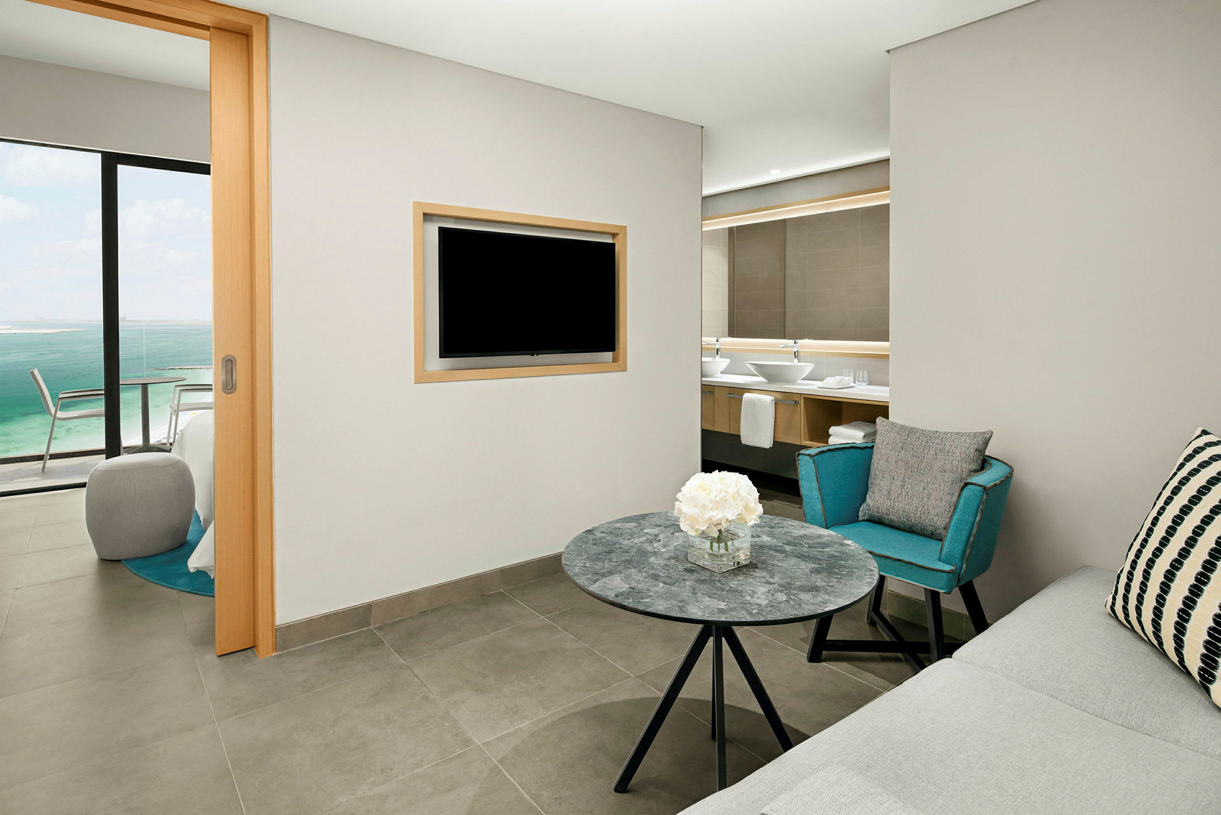 Ett hotellrum med sittgrupp, ett bord med blommor, en soffa, en fåtölj, och en TV monterad på väggen. I bakgrunden syns ett badrum och genom en glasskjutdörr en utsikt över havet. (AI-genererad text)