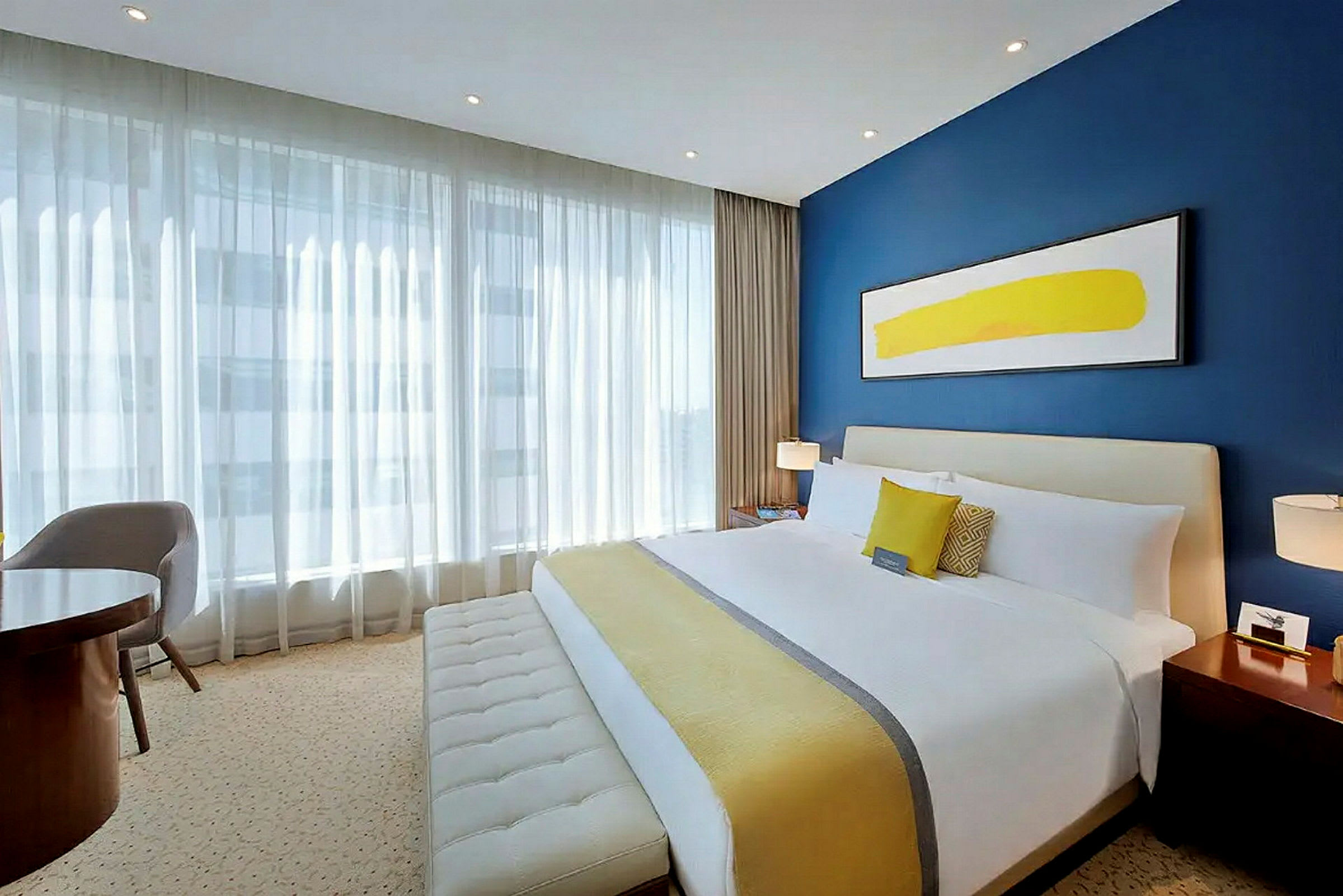 Ett hotellrum med en stor säng, blå vägg, modern konst och stora fönster med vit gardin. (AI-genererad text)